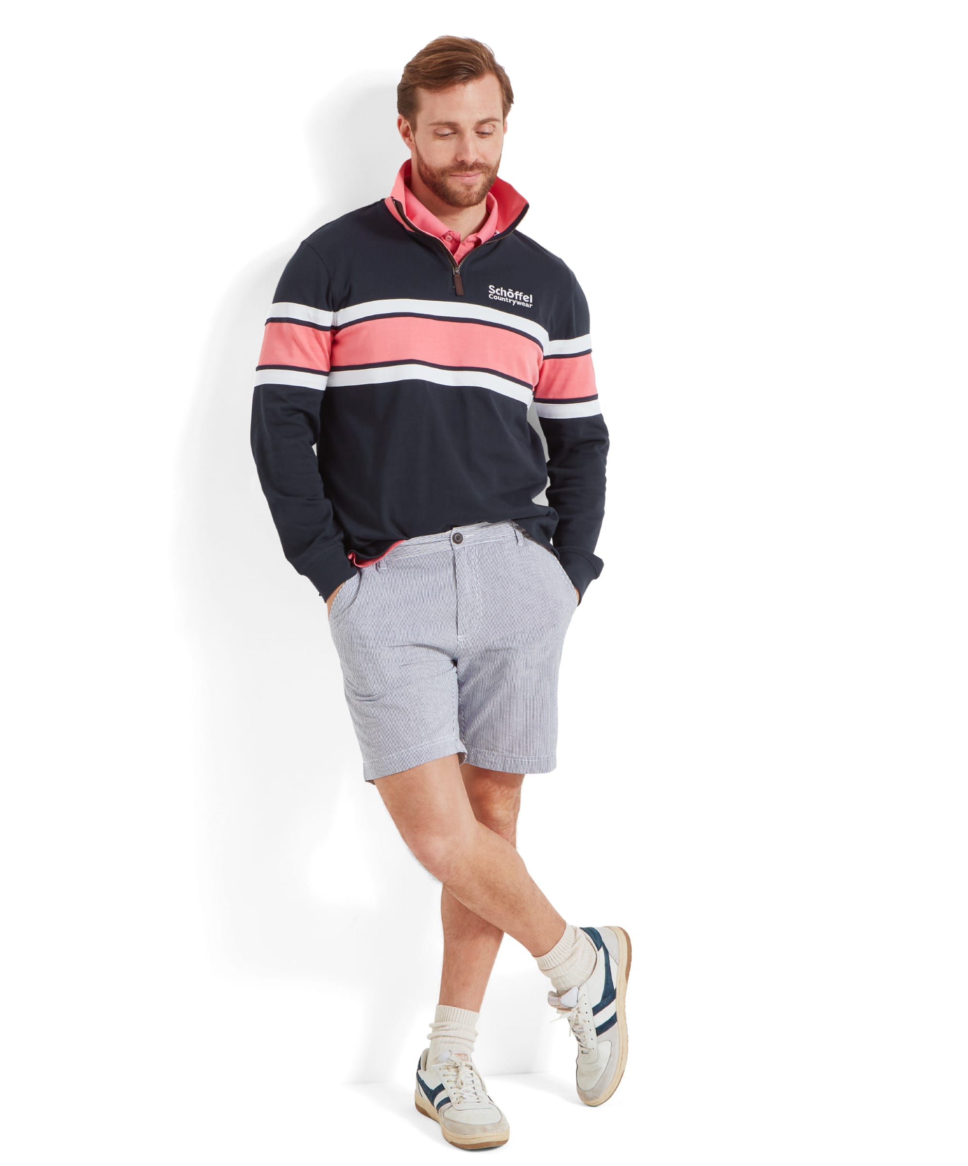 Full body shot of the man wearing the Schöffel Exmouth Heritage 1/4 Zip for Unisex in Navy, light blue striped shorts, and white trainers. He is standing with one leg crossed over the other, looking down and smiling slightly.