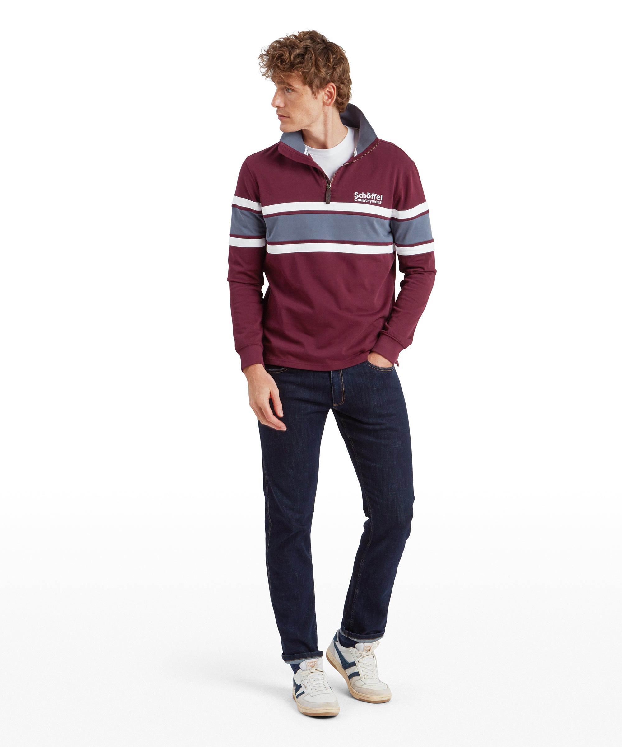 The same man is standing with his back slightly turned towards the camera. He is wearing the same Schöffel Exmouth Heritage 1/4 Zip for Unisex in Red, showing the back design, which includes blue and white horizontal stripes that continue across the back. He is also wearing dark blue jeans.