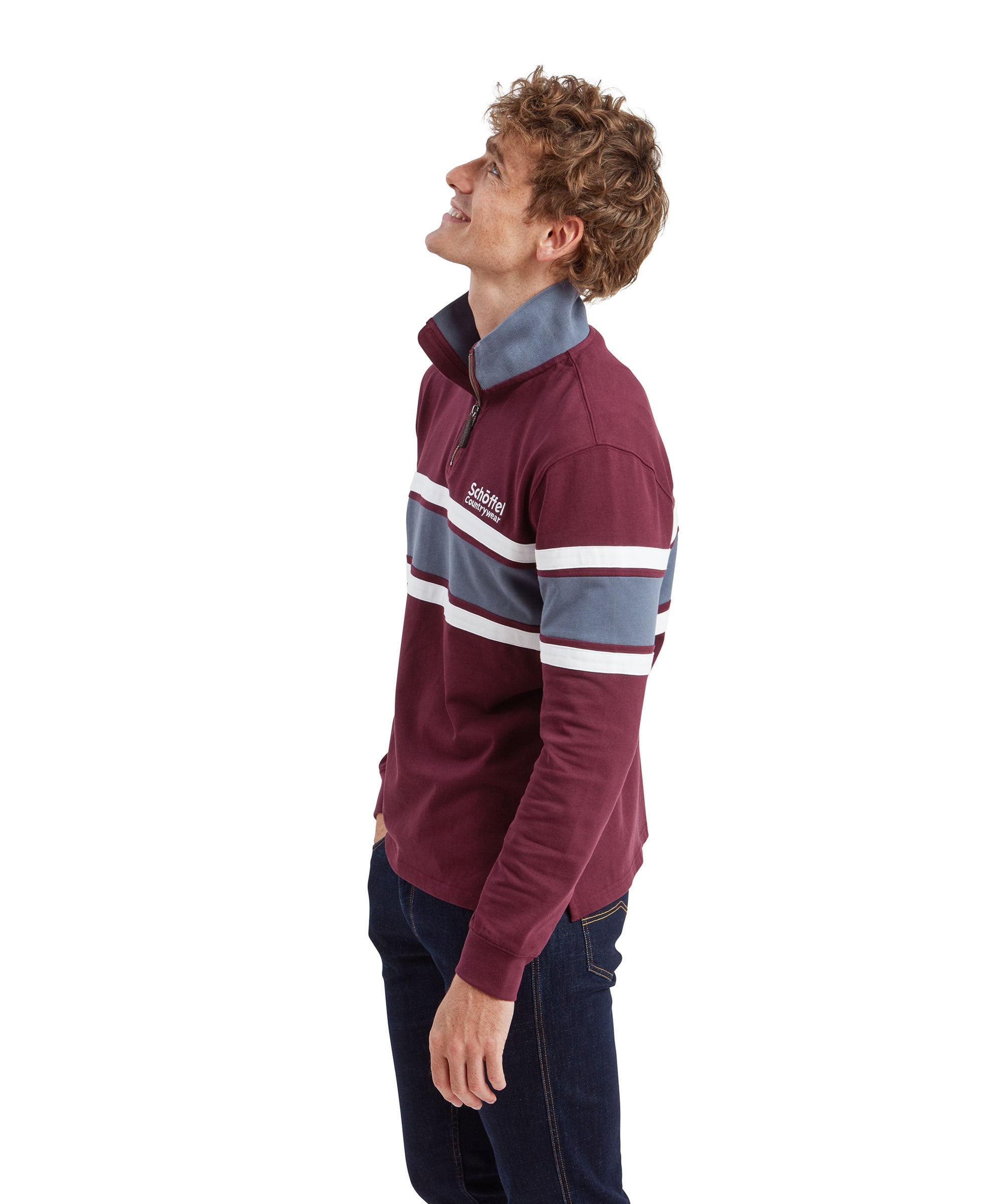 The man is standing and looking to the side with his hands in his pockets. He is wearing the Schöffel Exmouth Heritage 1/4 Zip for Unisex in Red with blue and white stripes and dark blue jeans. The setting is minimalistic with a plain white background.