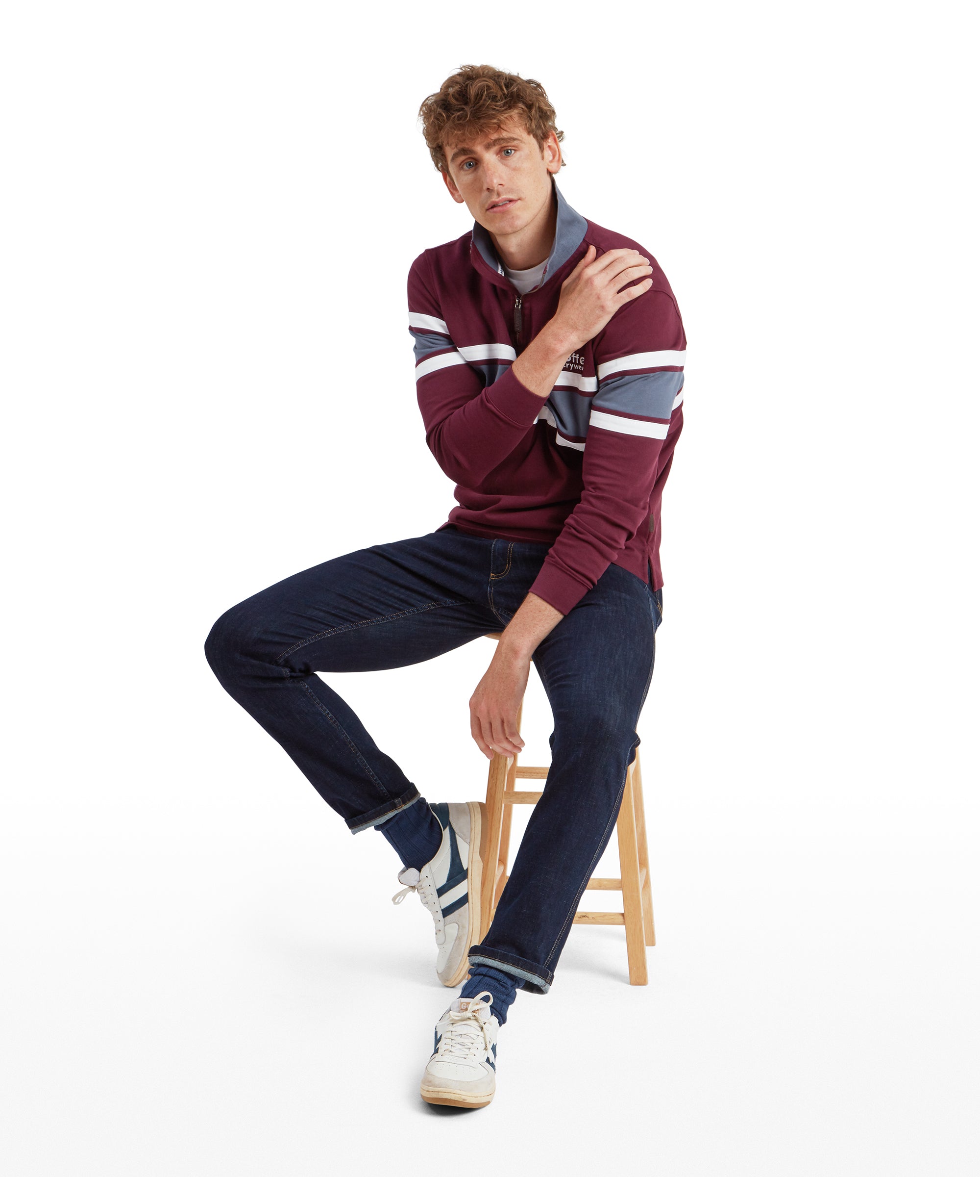 The man is sitting on a wooden stool, looking relaxed with one hand resting on his shoulder. He is wearing the Schöffel Exmouth Heritage 1/4 Zip for Unisex in Red with blue and white stripes and dark blue jeans. The outfit is casual and sporty.