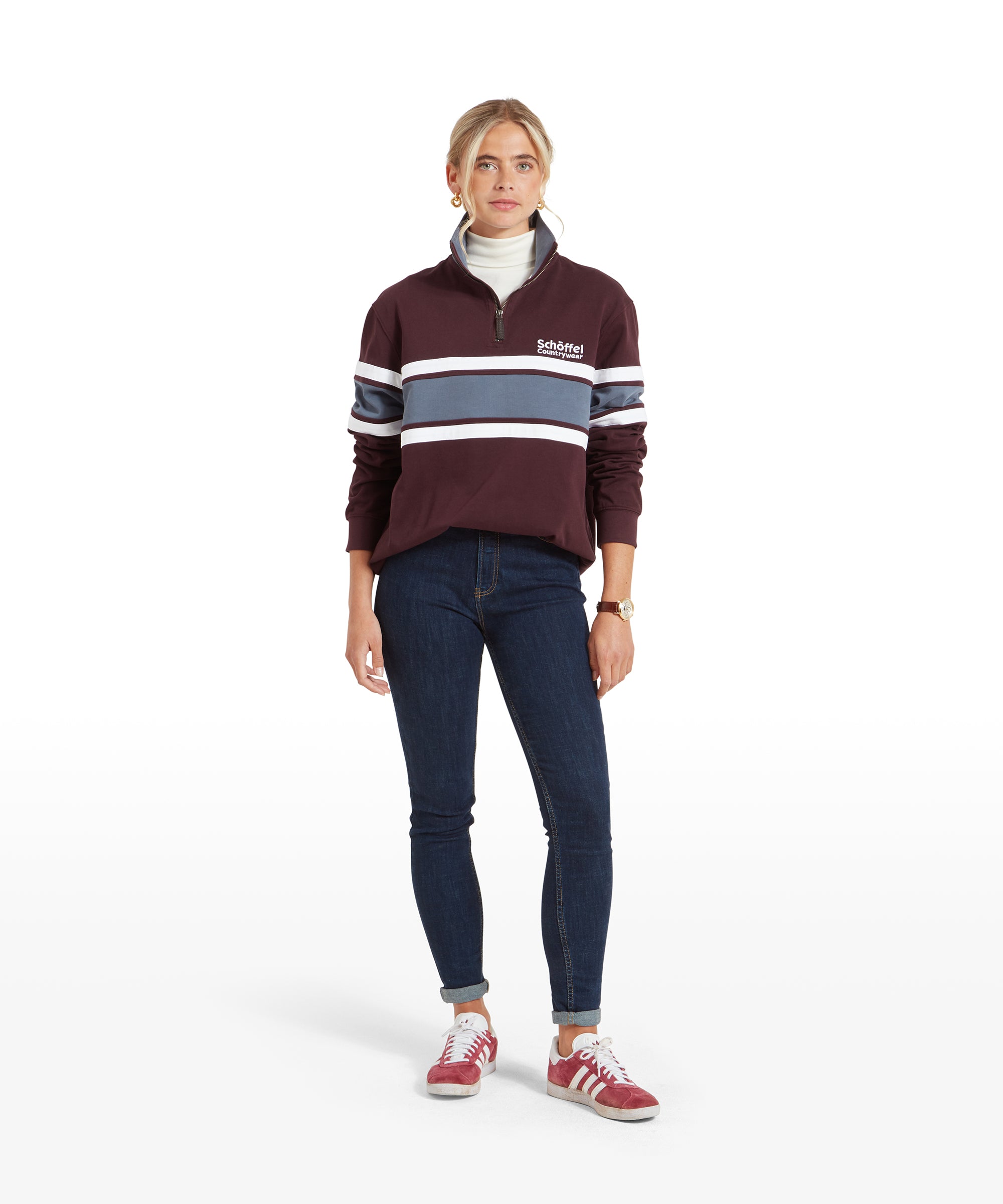 The same woman standing in a relaxed pose, showcasing the full outfit. She is wearing a Schöffel Exmouth Heritage 1/4 Zip for Unisex in Red with white and blue stripes, dark skinny jeans, and red trainers. The pullover has a high collar, and her hair is tied back.