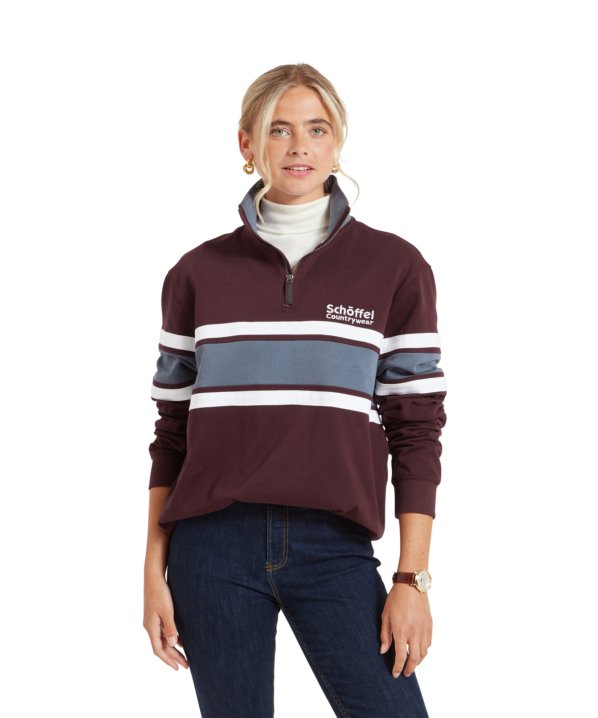 A woman is wearing a Schöffel Exmouth Heritage 1/4 Zip for Unisex in Red with white and blue horizontal stripes across the chest and arms. The pullover features the "Schöffel Countrywear" logo on the left side of the chest. She pairs it with dark blue jeans, a white turtleneck underneath, gold hoop earrings, and a wristwatch. Her blonde hair is styled back, and she is looking confidently forward.