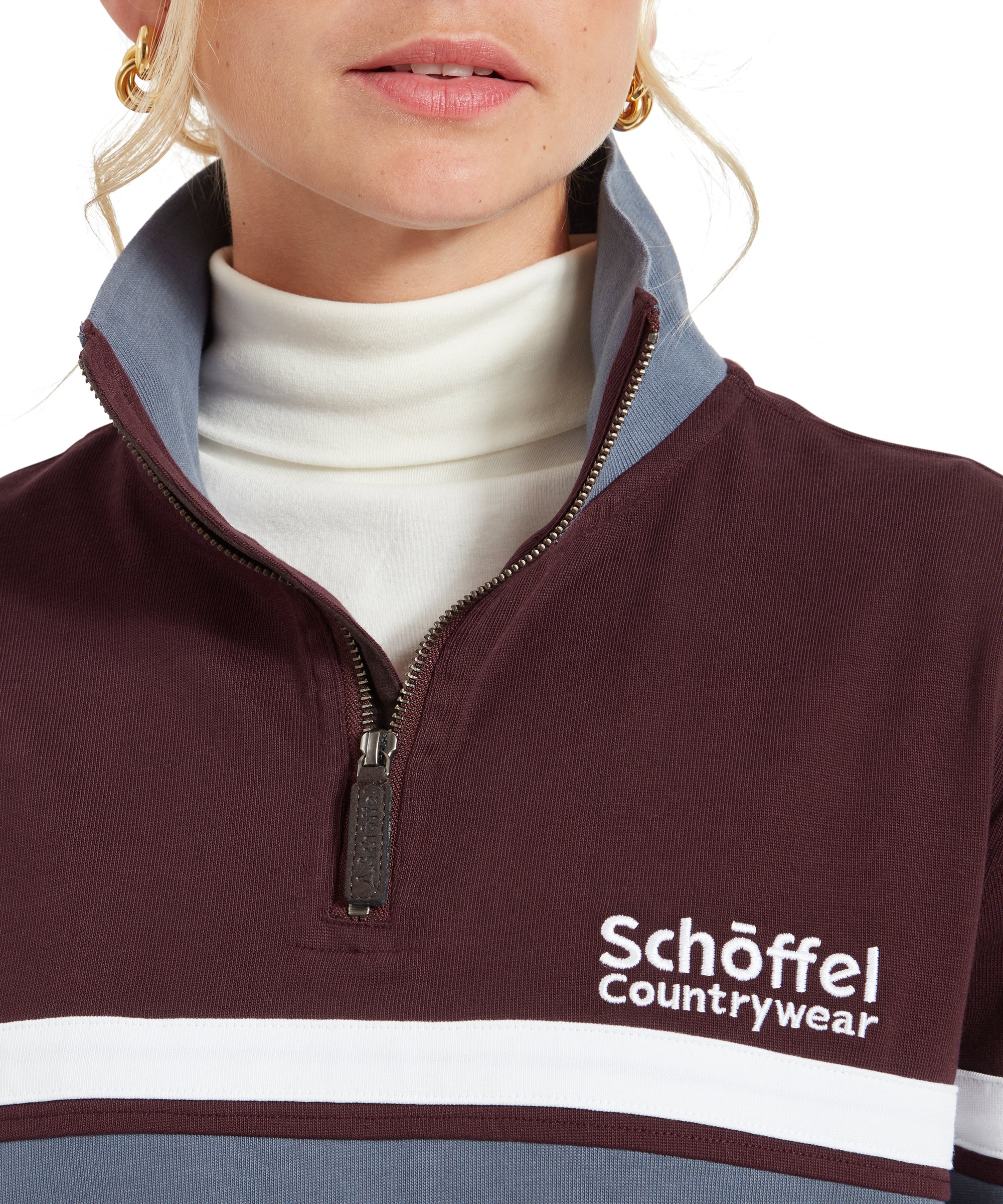 A close-up shot of the collar and zipper area of the Schöffel Exmouth Heritage 1/4 Zip for Unisex in Red, featuring the "Schöffel Countrywear" logo and a blue contrast collar over a white turtleneck. The zipper is halfway up, revealing the texture and detail of the fabric.