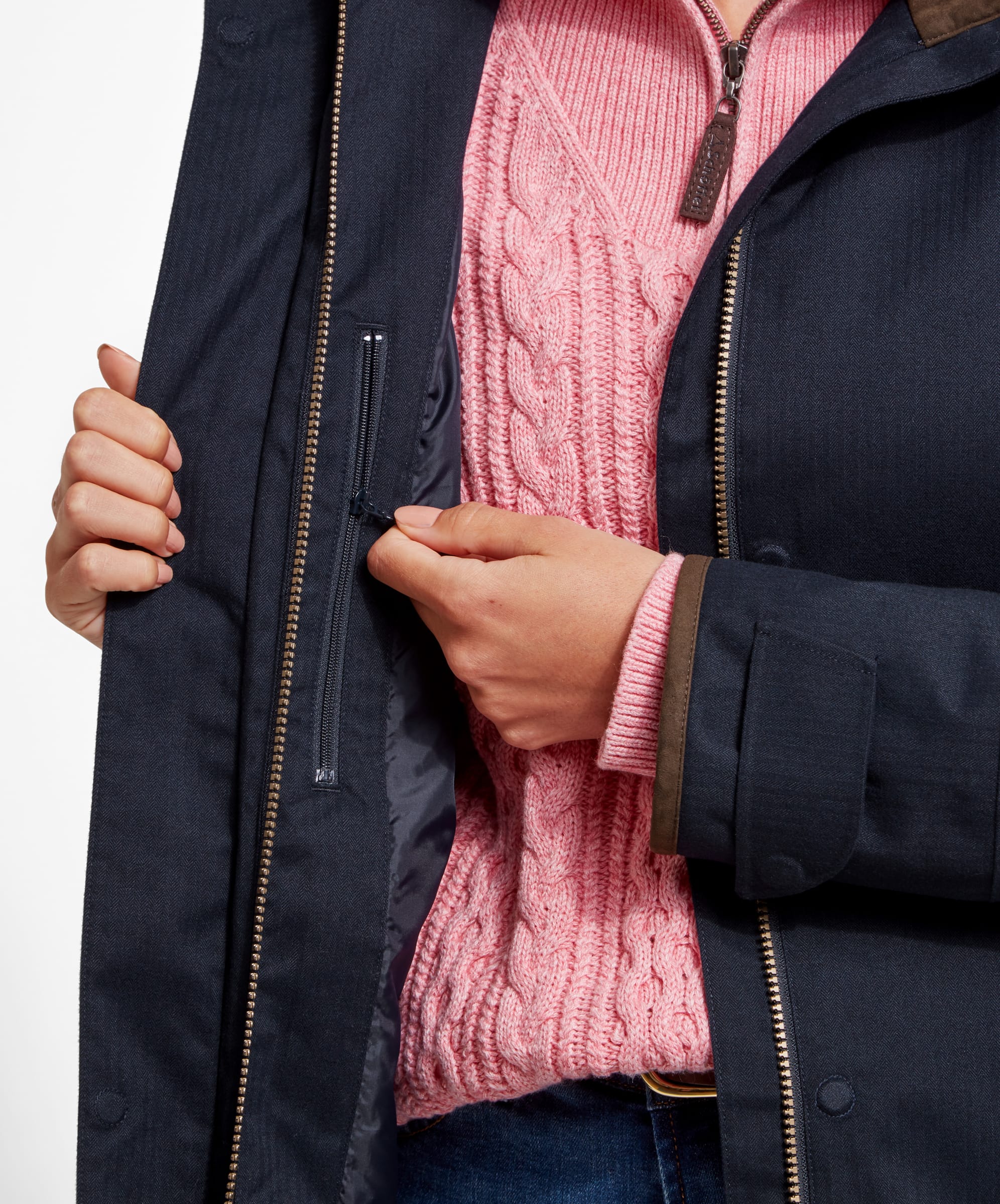 A close-up of a woman wearing a Schöffel Wakerley Jacket for Women in Blue with the front unzipped, revealing a pink cable-knit sweater underneath. The woman is holding the inner zipper of the jacket with one hand, showcasing the interior lining and an inner pocket of the jacket. The detailed texture of the pink sweater contrasts with the smooth, dark fabric of the jacket.