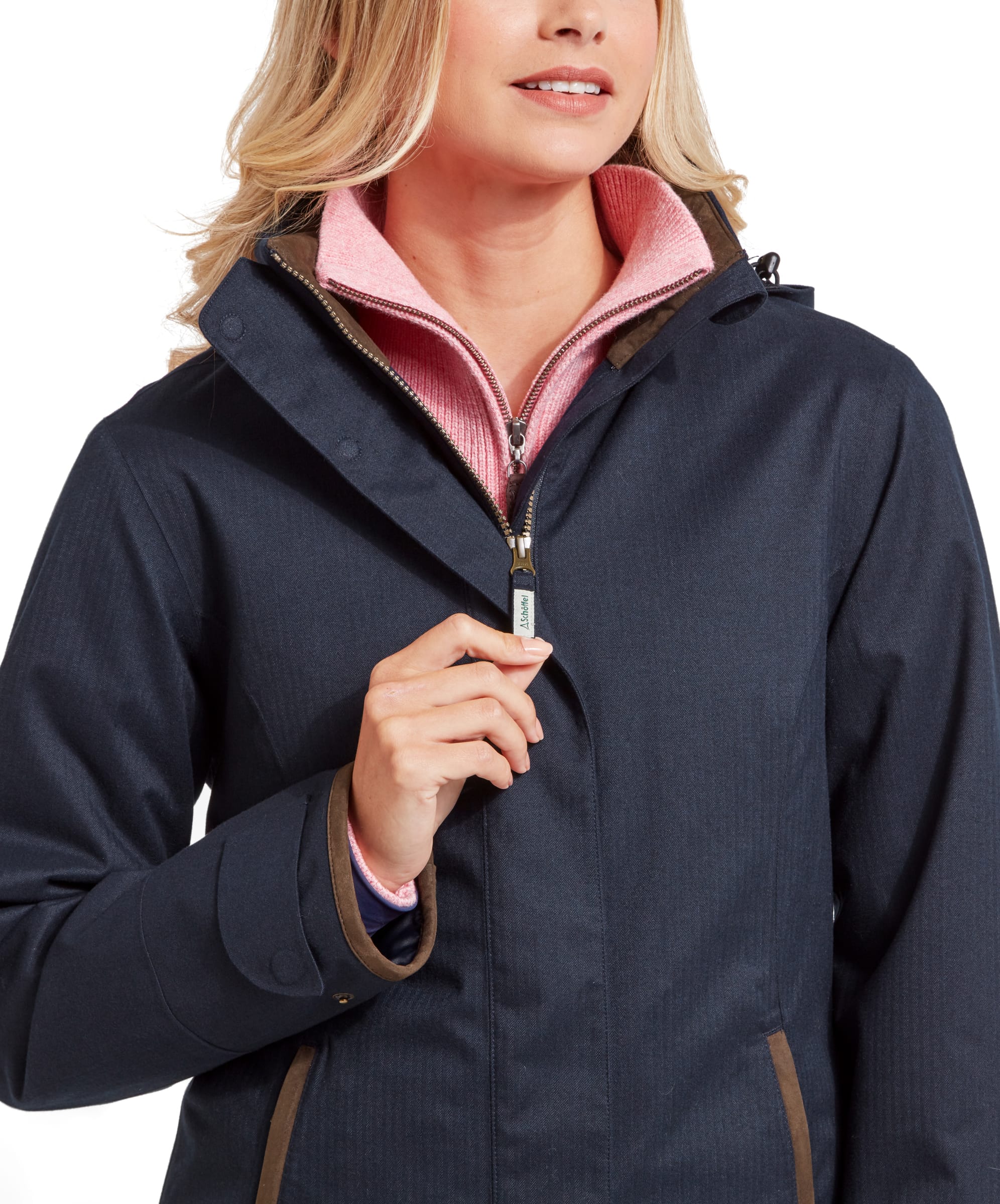 The woman zips up the front of her Schöffel Wakerley Jacket for Women in Blue. The pink sweater collar is visible underneath the jacket, providing a pop of colour. The jacket has a sleek design with brown accents on the collar and around the zipper.