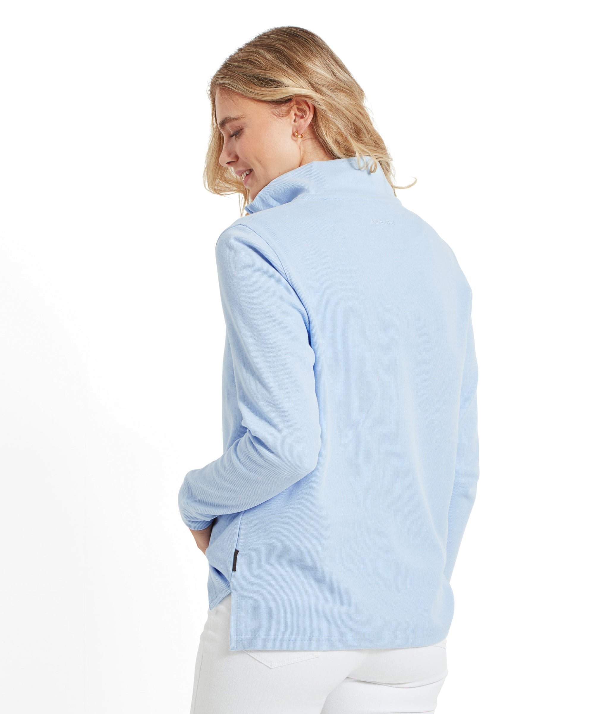 A model wearing a Schoffel women's Appletree Bay 1/4 zip jumper in blue, paired with white jeans, viewed from the back. She is smiling and looking down, with her blonde hair styled in loose waves.