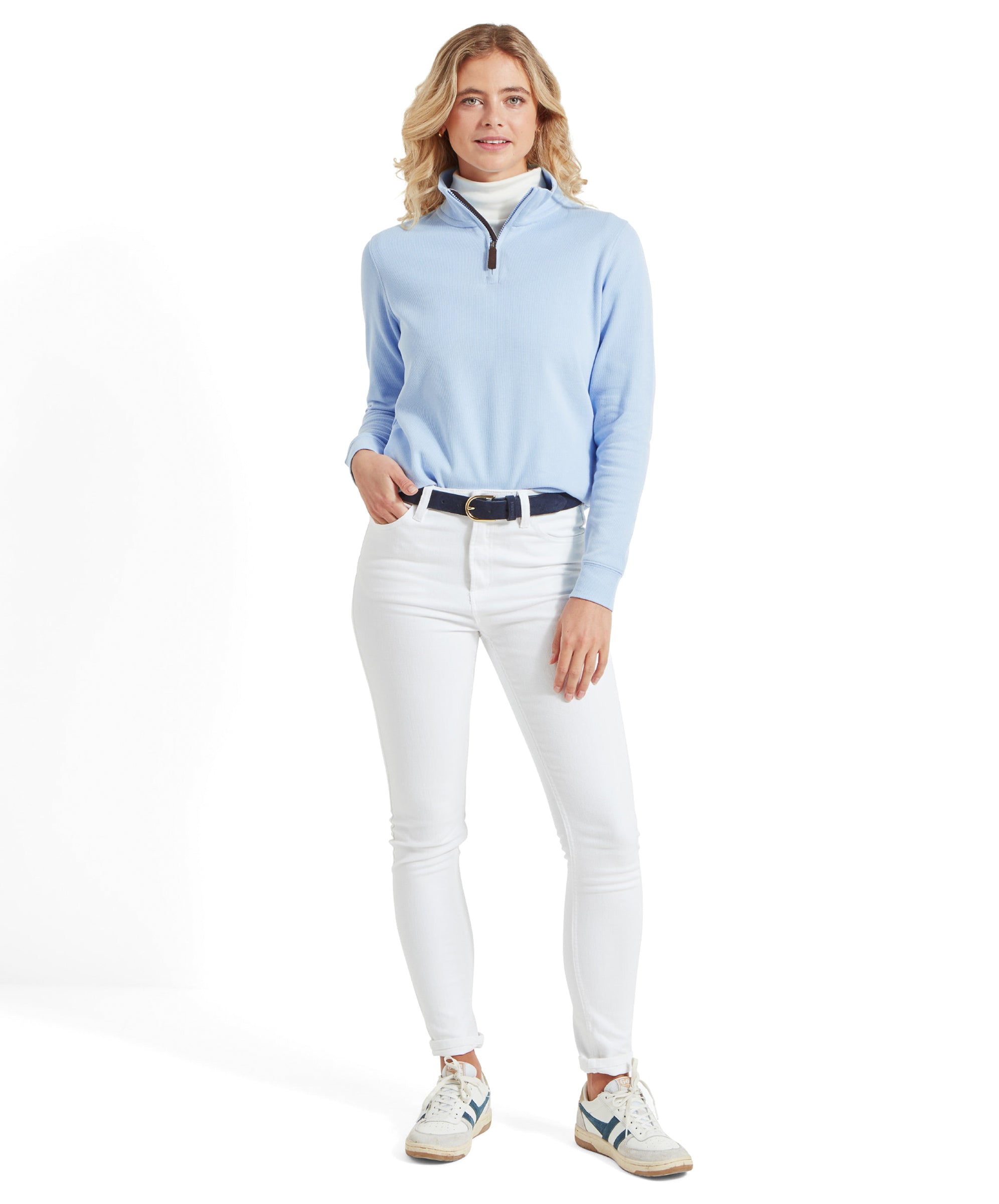 A model wearing a Schoffel women's Appletree Bay 1/4 zip jumper in blue, white turtleneck, white jeans, and white trainers with blue accents, standing and smiling.