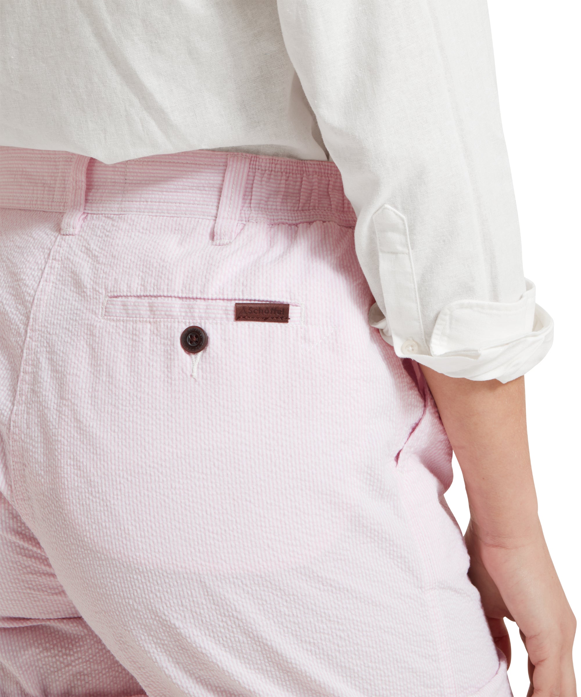 A close-up of a female model wearing pink and white stripe Schöffel shorts paired with a white tucked in shirt. She is standing in a relaxed pose with one hand in the pocket. The image highlights the shorts elasticated waist back pocket