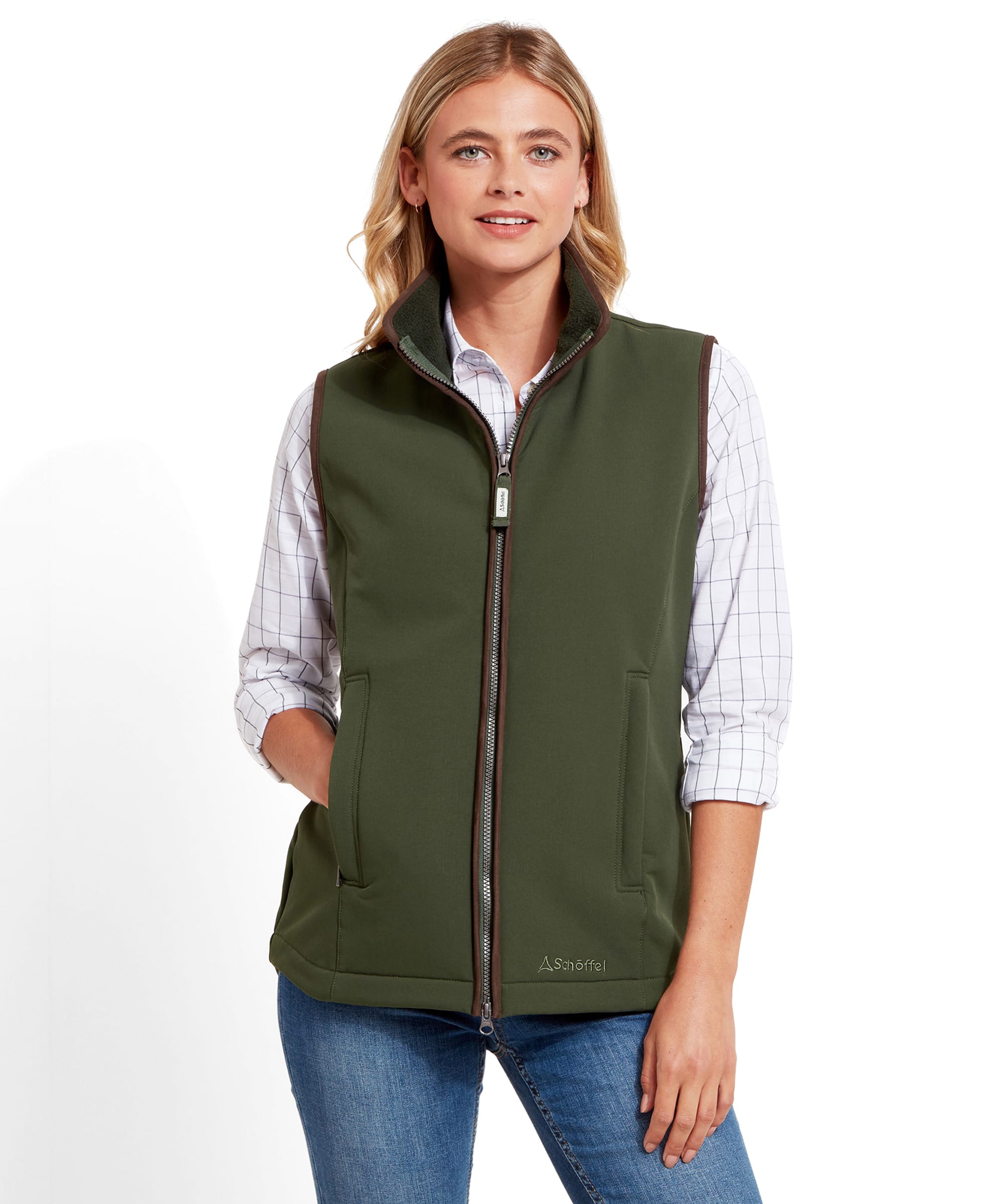 A woman is wearing a Schöffel Belton Gilet for Women in Green over a checkered shirt, standing with her hands in her pockets, smiling. The gilet features a full zip front and a high collar.