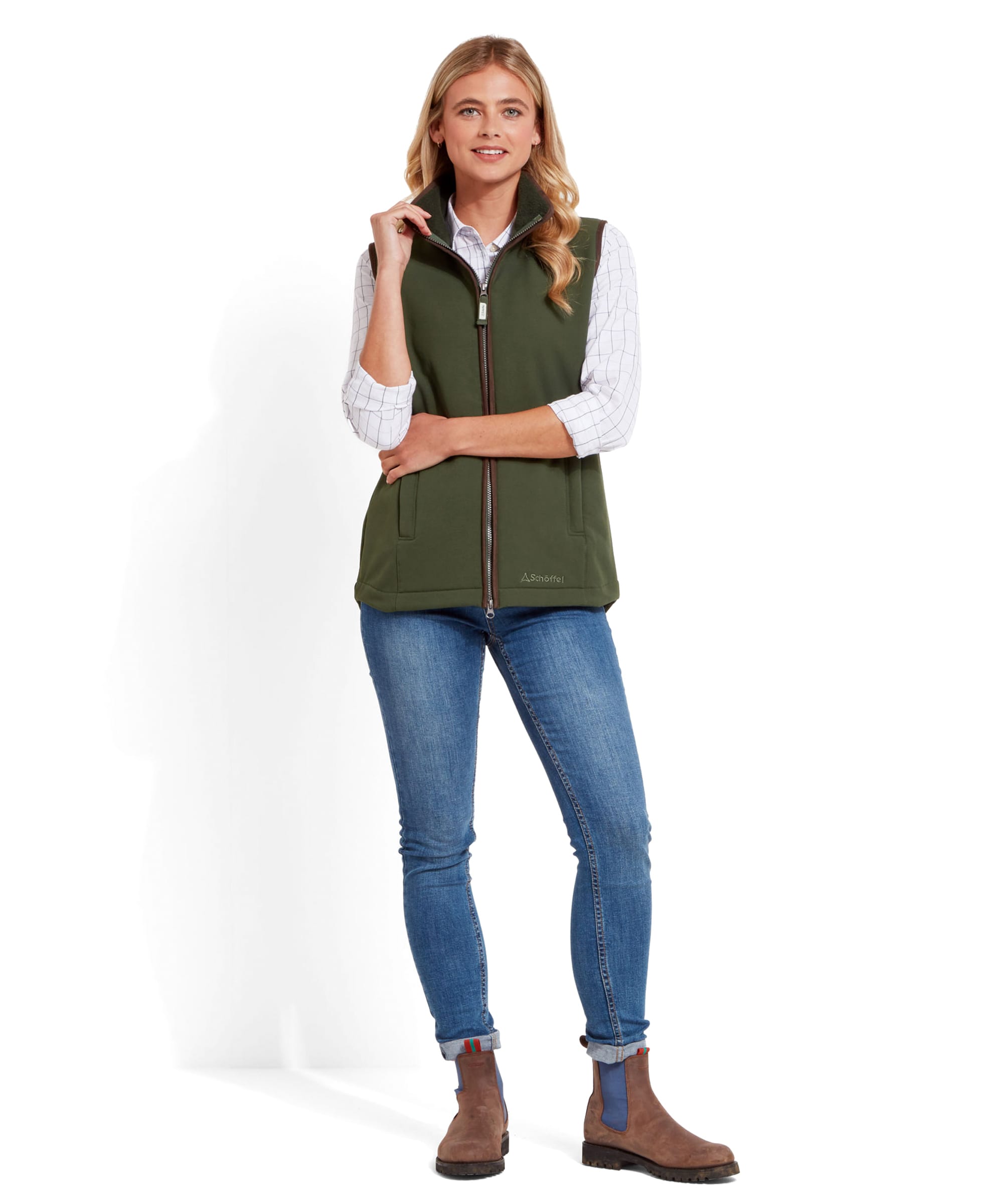 The same woman is wearing a Schöffel Belton Gilet for Women in Green over a checkered shirt, paired with blue jeans and brown ankle boots. She is standing with one hand in her pocket and the other hand lightly touching her collar.