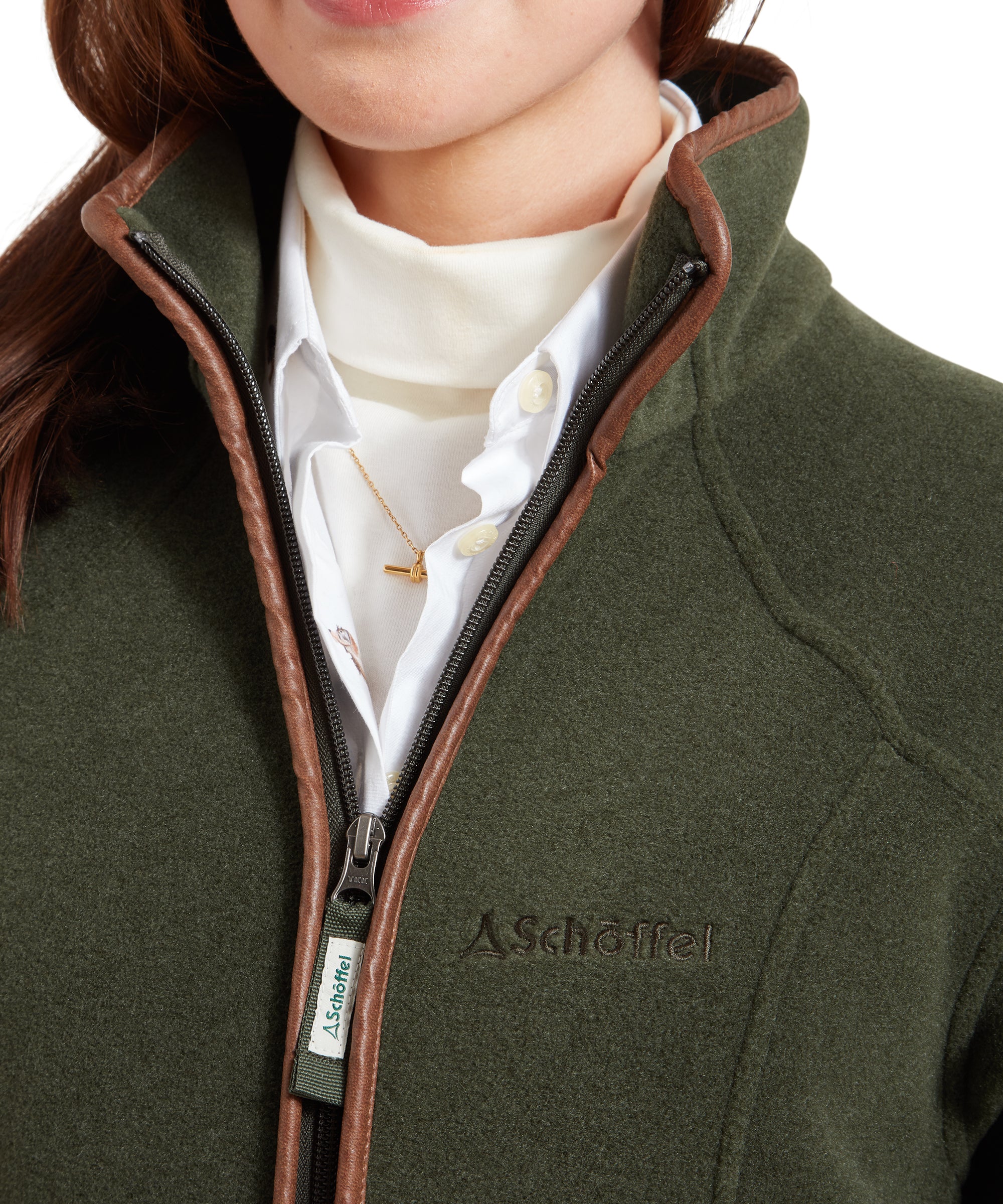 A close-up of the Schöffel Burley Fleece Jacket for Women in Green's zipper area. The Schöffel logo is embroidered on the chest, and the zipper is detailed with a leather trim. The woman is also wearing a white shirt underneath.