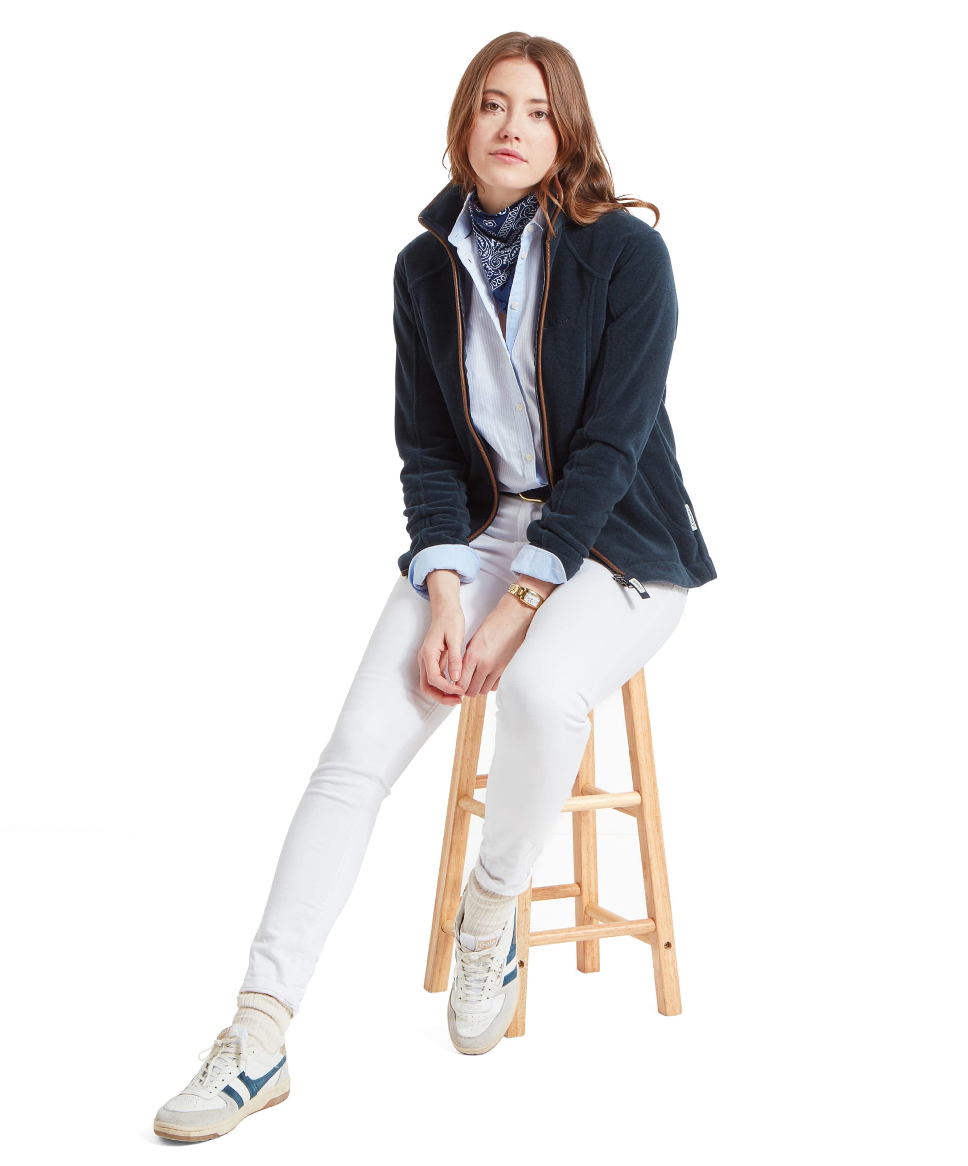 The same woman is seated on a wooden stool, wearing the Schöffel Burley Fleece Jacket for Women in Blue, white jeans, and casual TRAINers, looking at the camera with a relaxed posture.