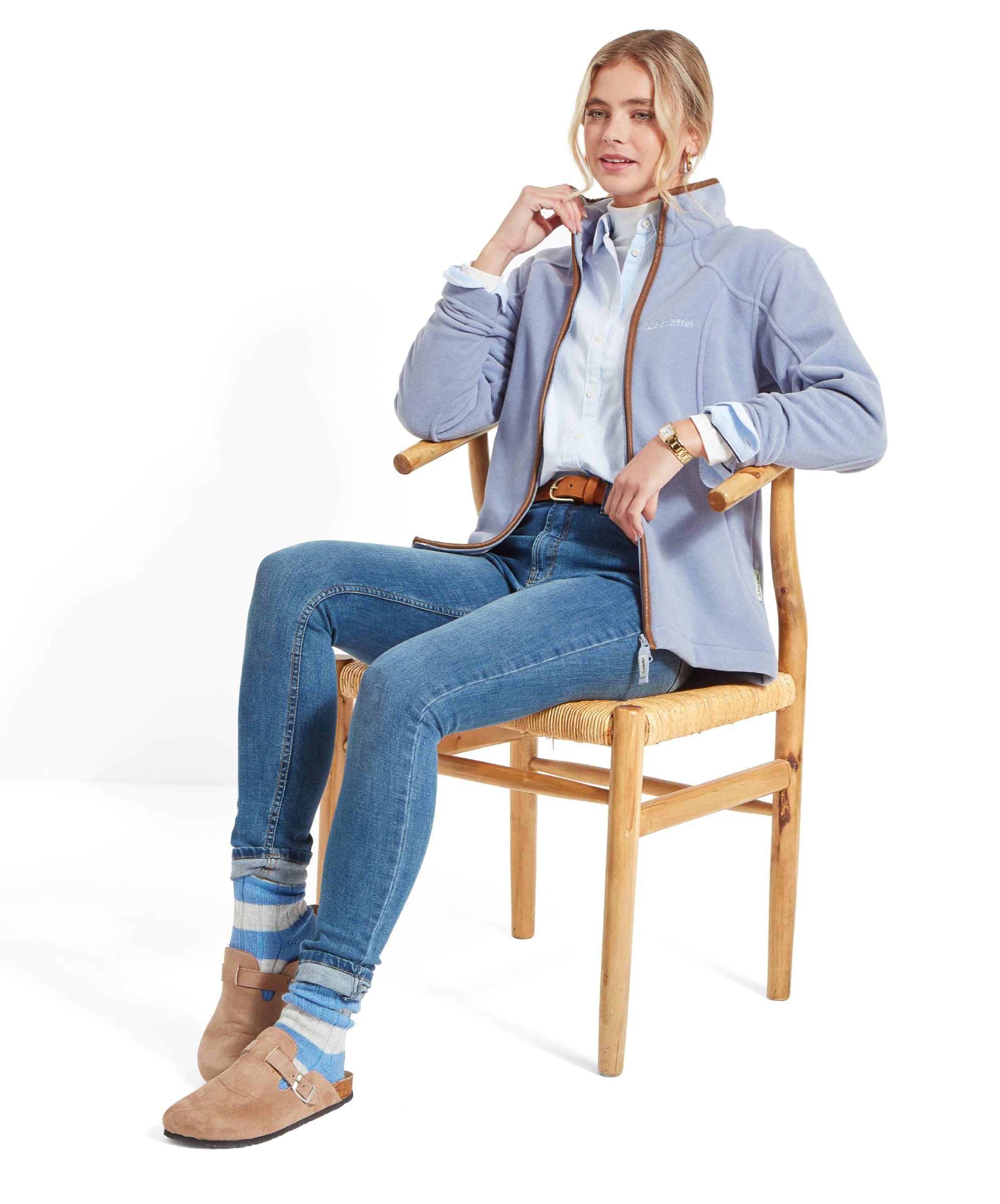 Woman sitting on a wooden chair, wearing a Schöffel Burley Fleece Jacket for Women in Light Blue, blue button-up shirt, and blue jeans. Her outfit is completed with light brown footwear and socks.