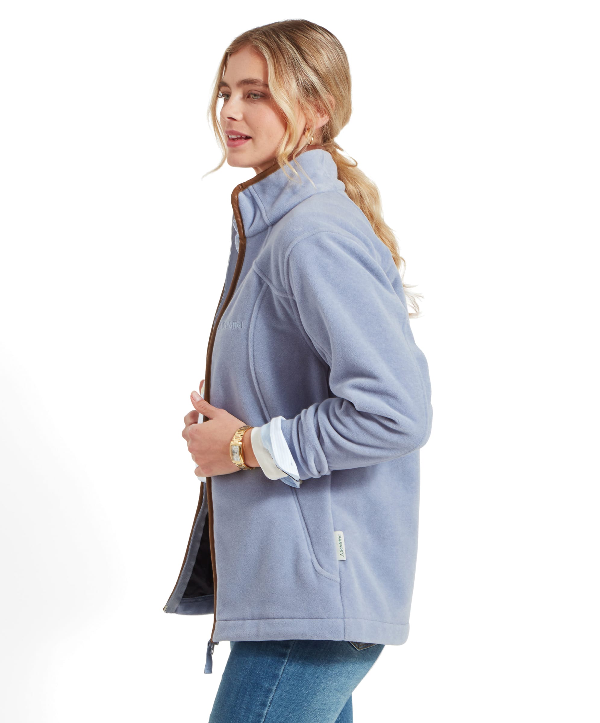 Side view of a woman wearing a Schöffel Burley Fleece Jacket for Women in Light Blue. She is looking slightly away from the camera, with her blonde hair tied back. The jacket has a zippered front and is paired with a blue button-up shirt and jeans.