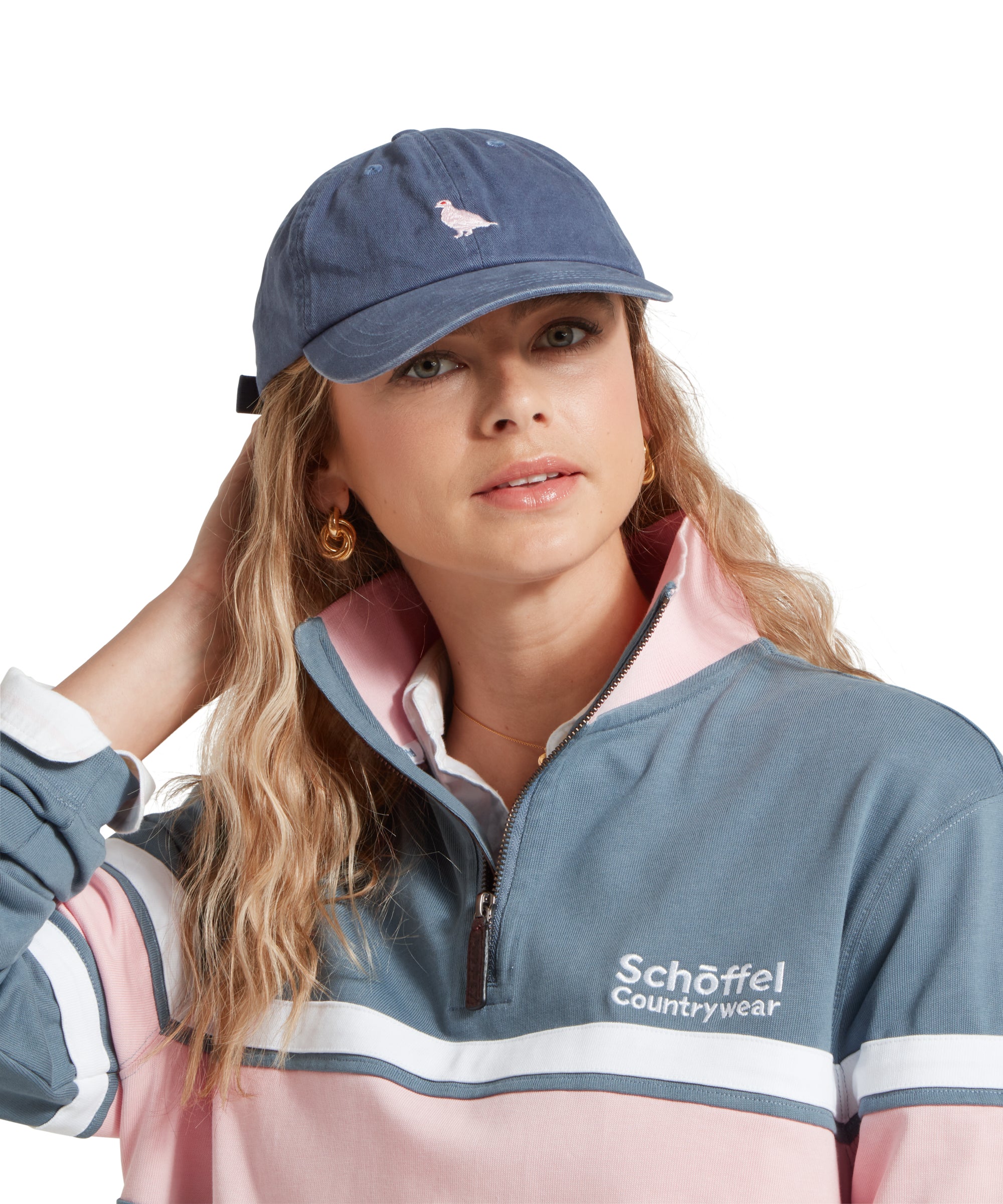 A close-up of a female model wearing a mid blue cap with white embroidered bird logo on the front. It is paired with a light blue and pink sweatshirt and white shirt.