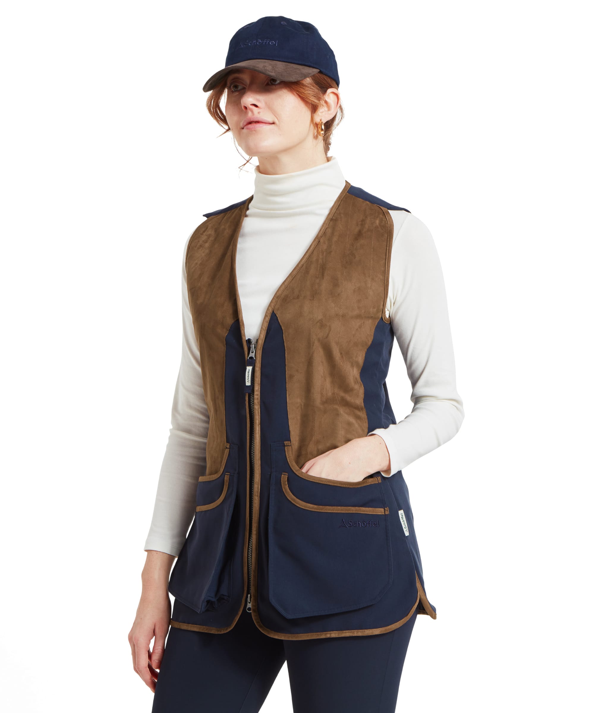 A woman wearing a Schöffel Edenham Clay Shooting Vest for Women in Blue over a white turtleneck and navy trousers. She also wears a navy cap and stands with her hands in the vest pockets.