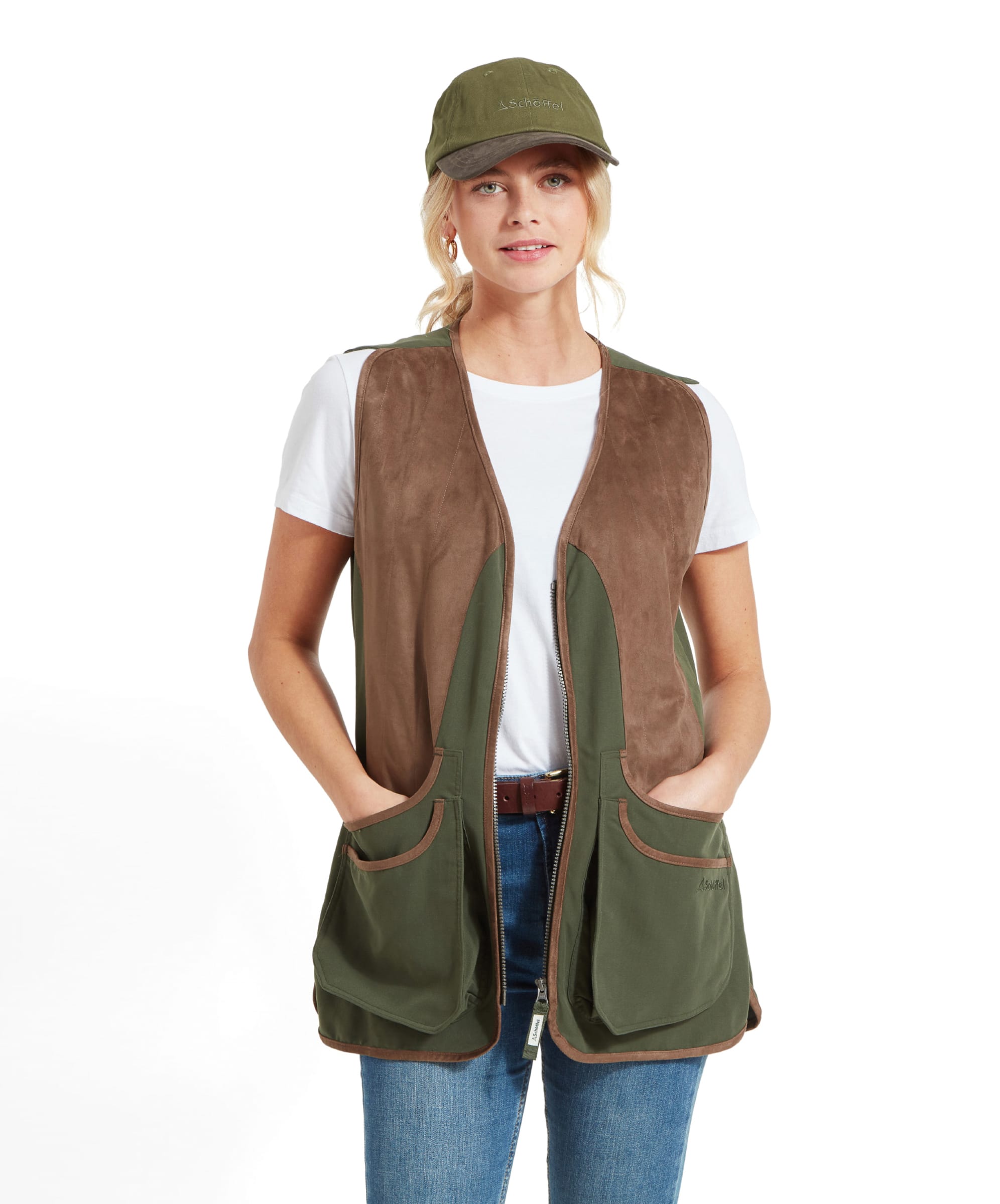 A woman is wearing a Schöffel Edenham Clay Shooting Vest for Women in Green over a white t-shirt, paired with a green cap. The vest features large front pockets and a zip-up front. She stands facing forward with her hands in the pockets, looking slightly to the side.