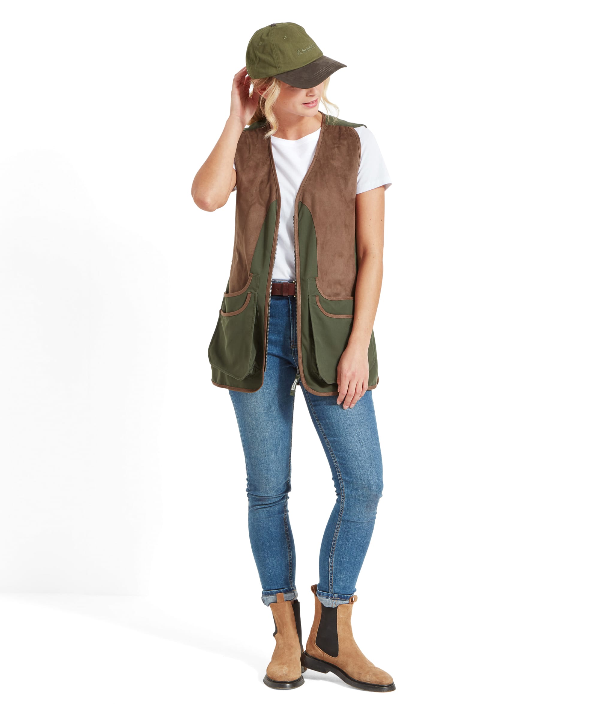 A woman is modeling a Schöffel Edenham Clay Shooting Vest for Women in Green, wearing a white t-shirt and blue jeans. She pairs the outfit with a green cap and tan ankle boots. She is standing in a casual pose, looking down while slightly adjusting her cap with one hand.