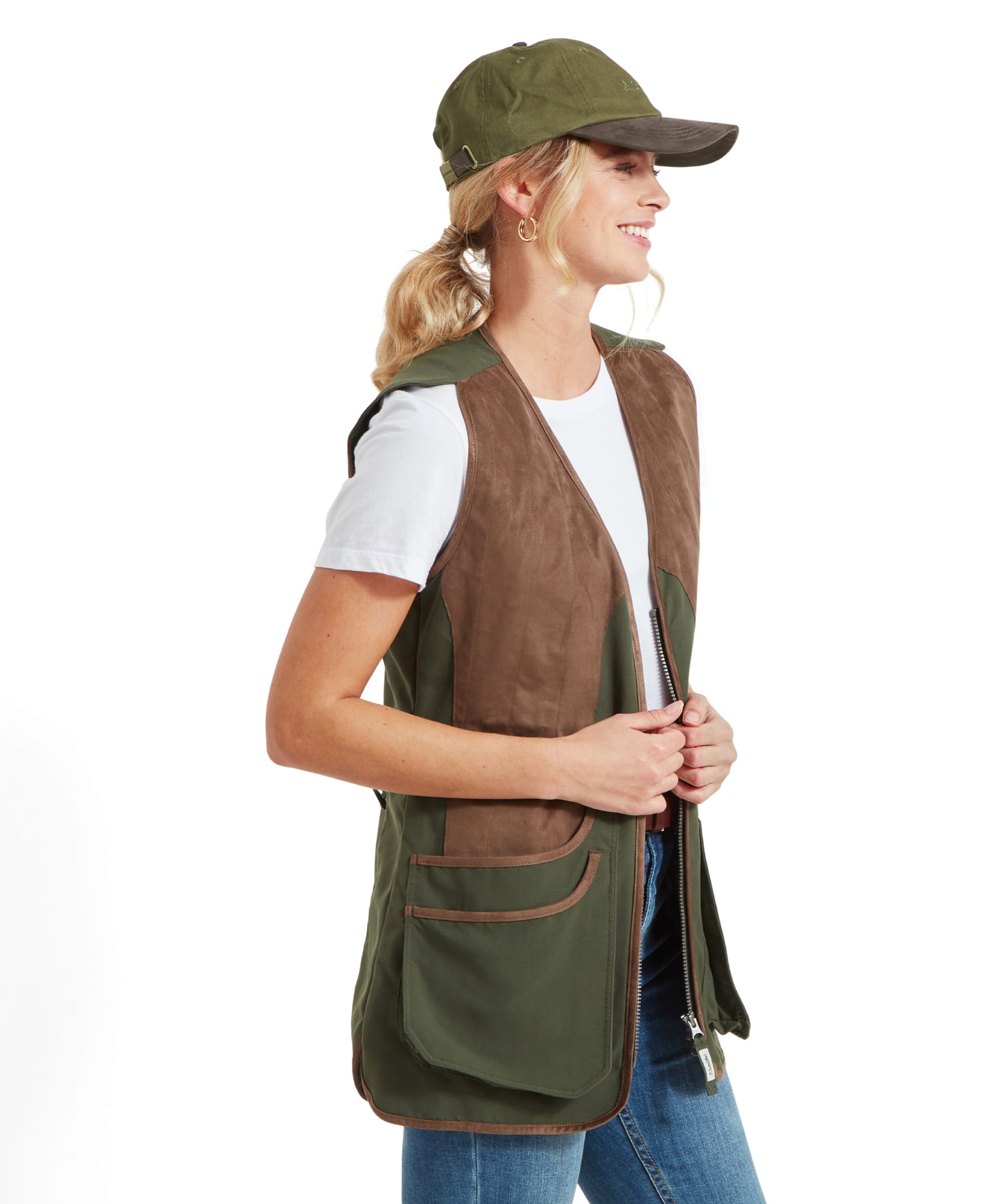 A side view of a woman wearing a Schöffel Edenham Clay Shooting Vest for Women in Green. She pairs it with a white t-shirt, blue jeans, and a green cap. She stands in profile, smiling and looking off to the side.