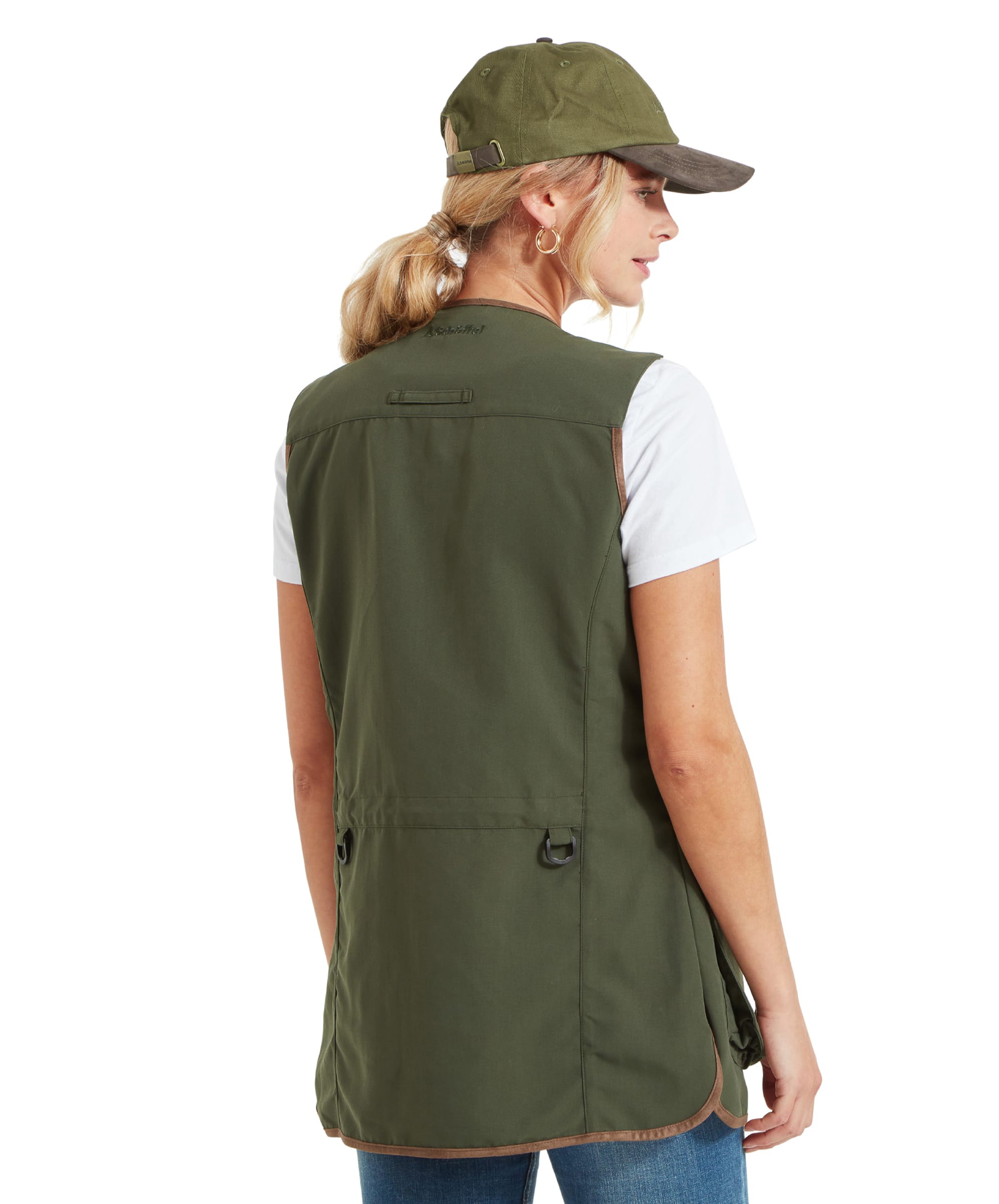 A woman is wearing a Schöffel Edenham Clay Shooting Vest for Women in Green with a white t-shirt and blue jeans. The image shows the back view of the vest, highlighting the drawstring detail at the waist and D-rings on the sides.