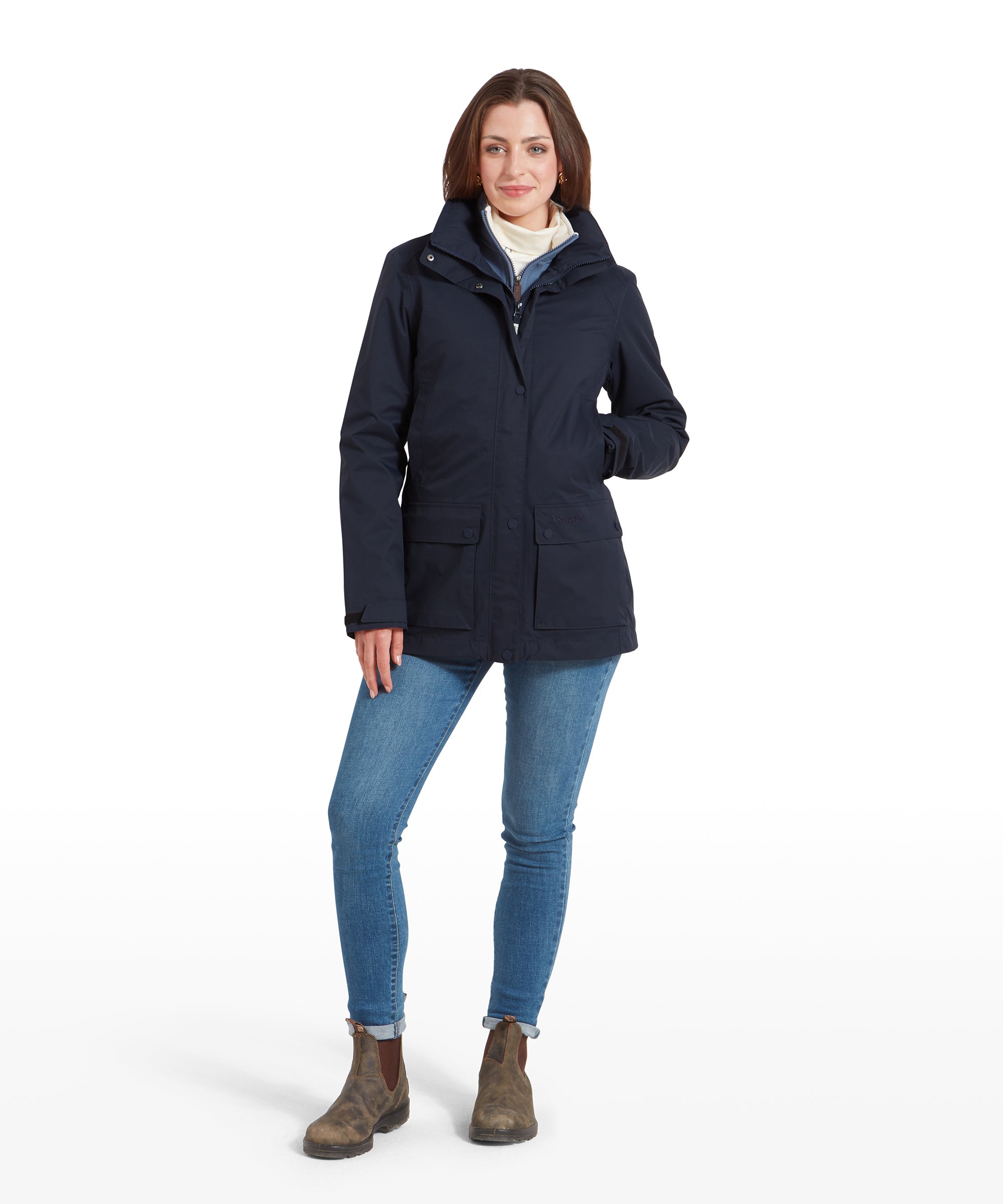 A woman is standing fully upright, wearing a Schöffel Edith Jacket for Women in Blue paired with blue jeans and brown boots. The jacket has a front zip and button closure, with a high collar and a fitted waist.