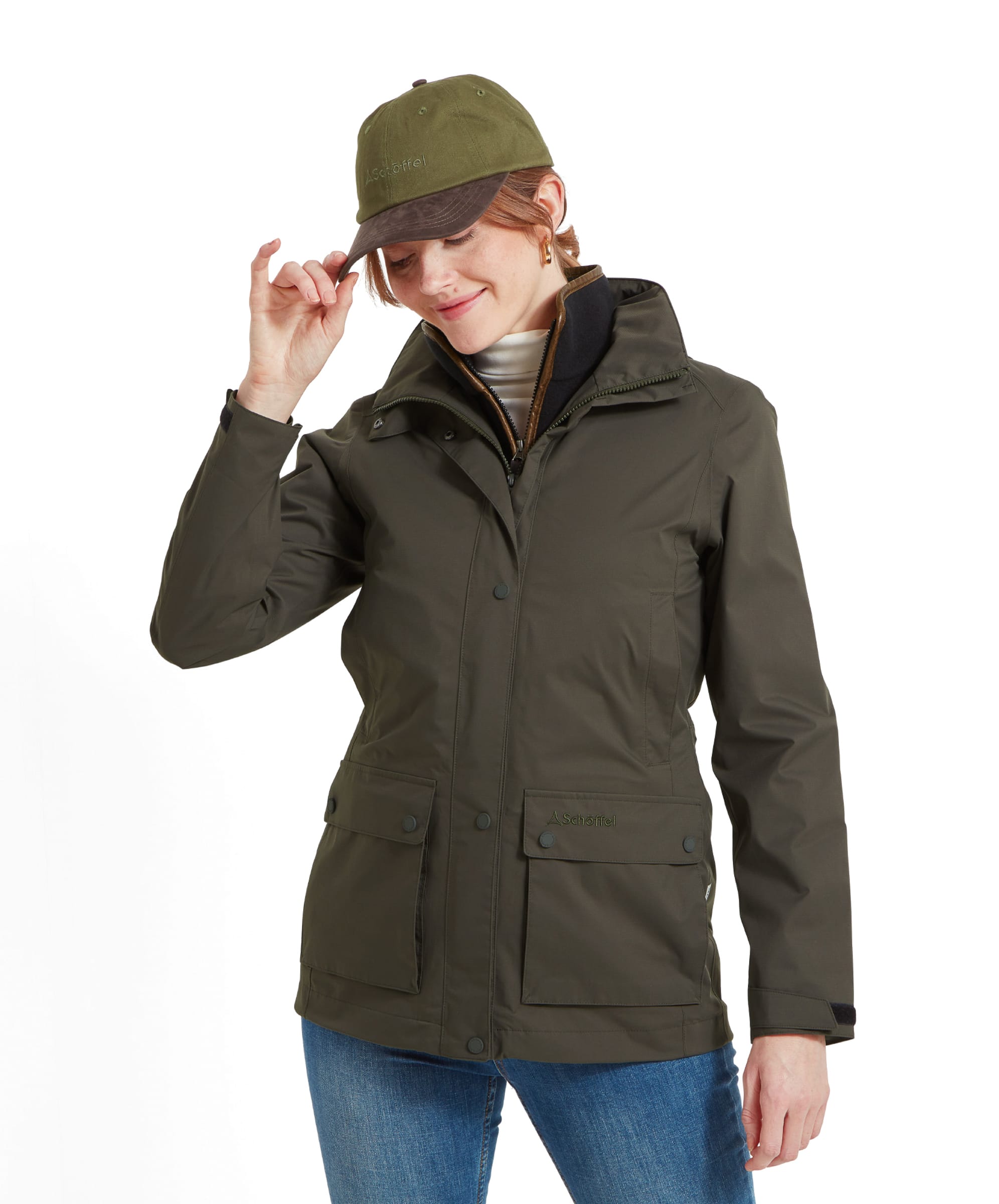 A woman wearing a Schöffel Edith Jacket for Women in Green, standing with her head slightly tilted, touching the brim of her green cap. The jacket has a front zipper and button closure with pockets.