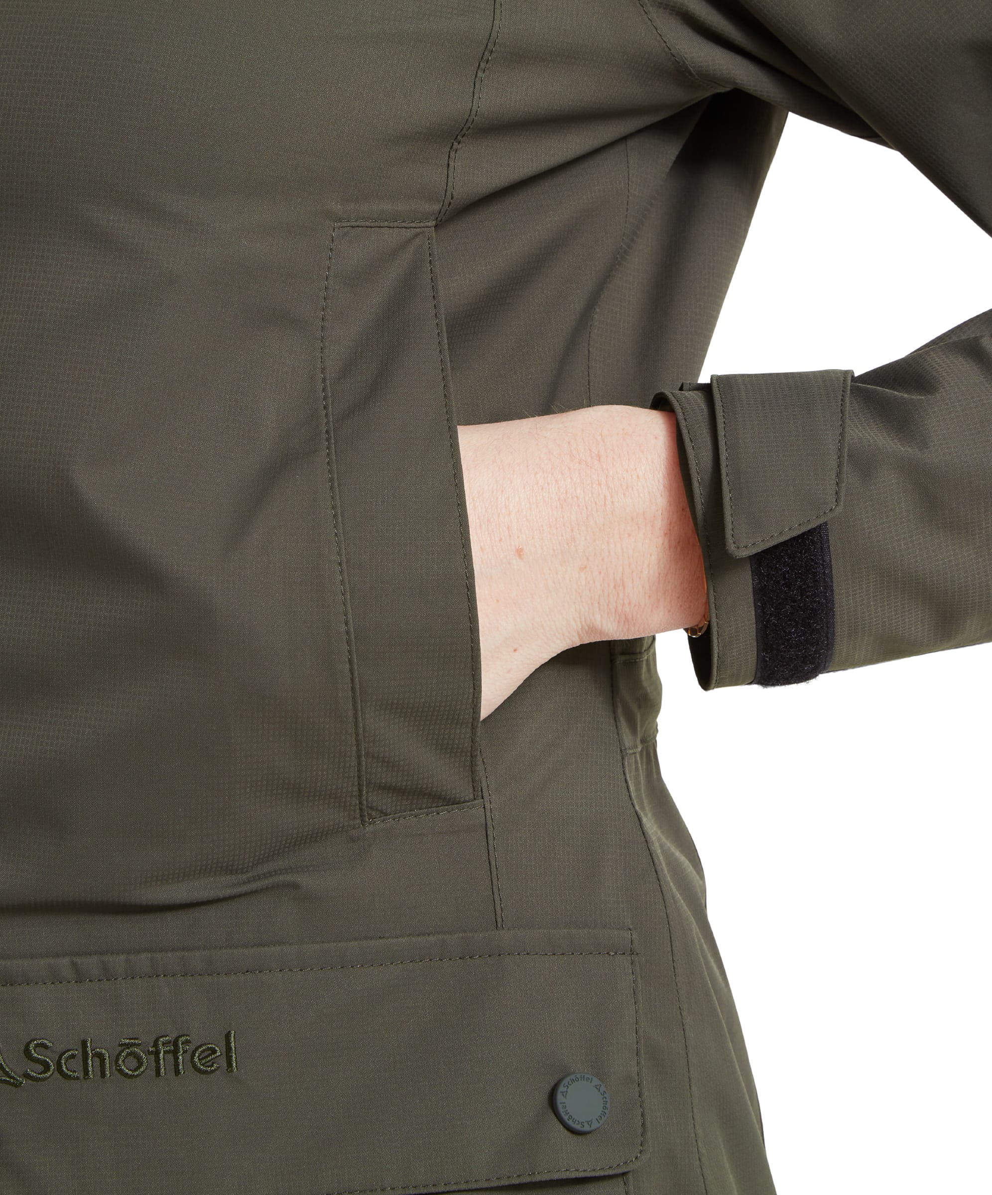 Close-up shot of a woman's hand placed inside the front pocket of an Schöffel Edith Jacket for Women in Green, showing the pocket flap and brand logo embroidery.