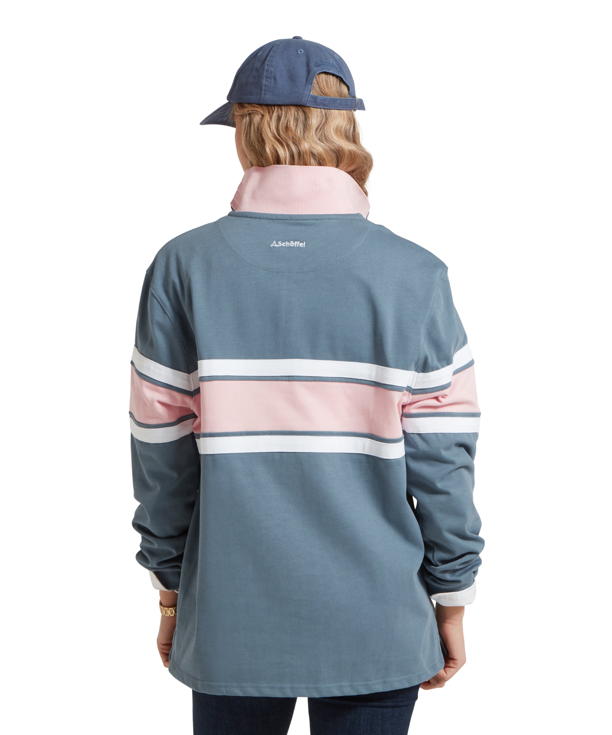 A back view image of a women wearing a blue Schöffel sweatshirt with pink and white stripe across the middle and small embroidered Schöffel logo. It is paired with a mid blue cap, white shirt and dark blue jeans and she is standing in a relaxed pose.