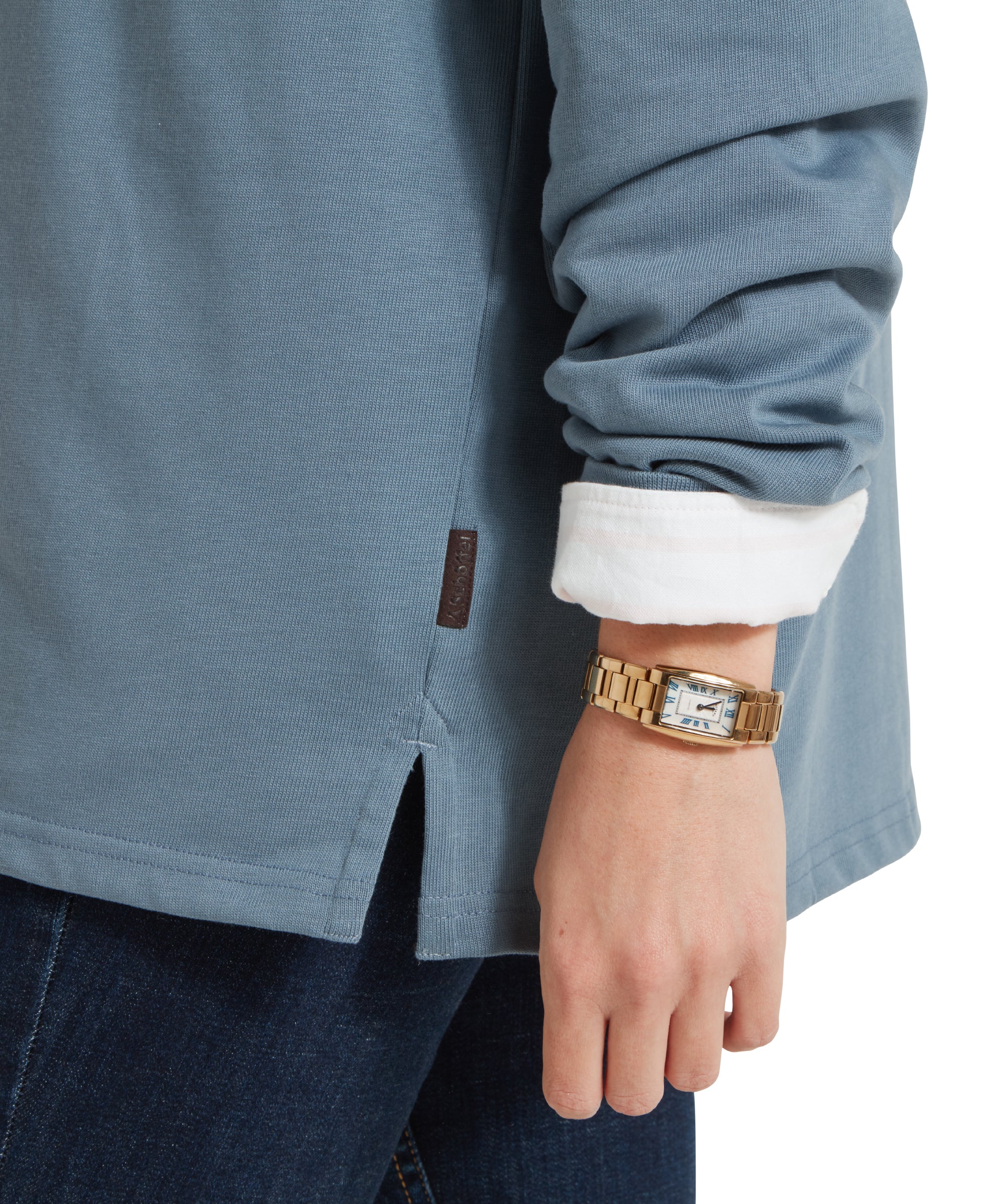 A close-up image of cuff and hem of a blue Schöffel sweatshirt with pink and white stripe across the middle and small embroidered Schöffel Countrywear logo. It is paired with a white shirt, dark denim jeans and gold jewellery.