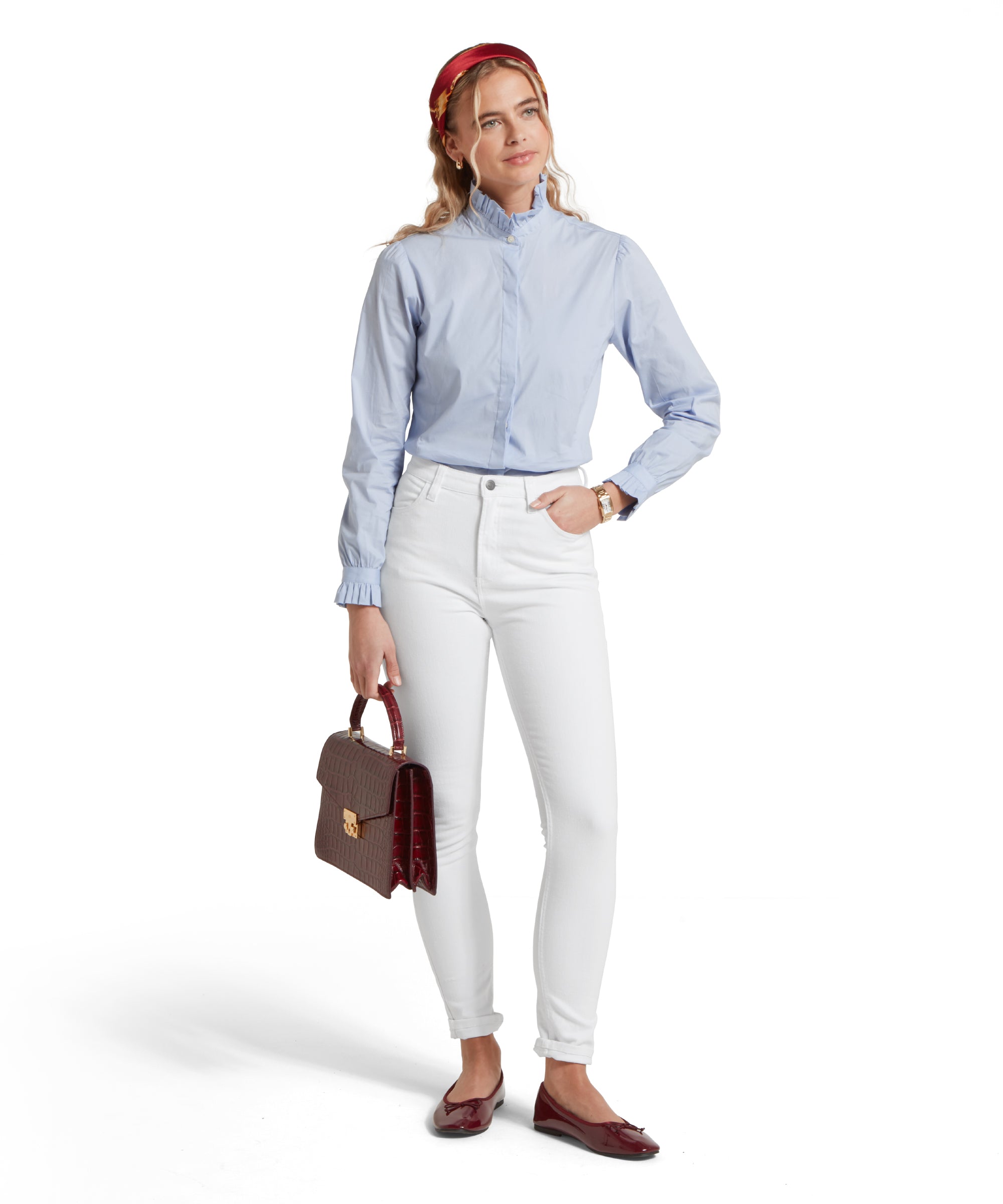 Full-height image of a female model wearing a light blue Schöffel shirt, paired with white jeans. She is standing in a relaxed pose with one hand in her pocket and a red leather handbag in the other.