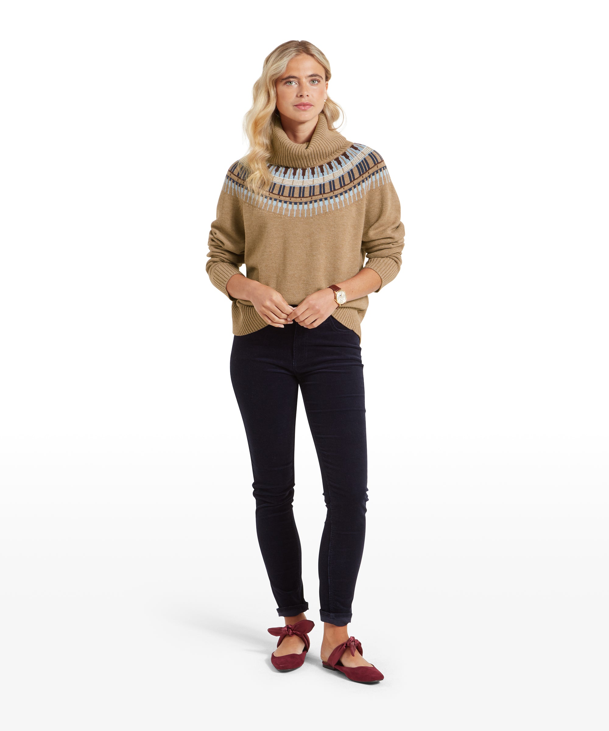 The same woman wearing the Schöffel Filey Fairisle Roll Neck Jumper for Women in Beige, shown in a full-body view. She is standing straight, with her hands lightly clasped in front of her, wearing dark fitted jeans and red flat shoes with bows. Her expression is calm and confident.