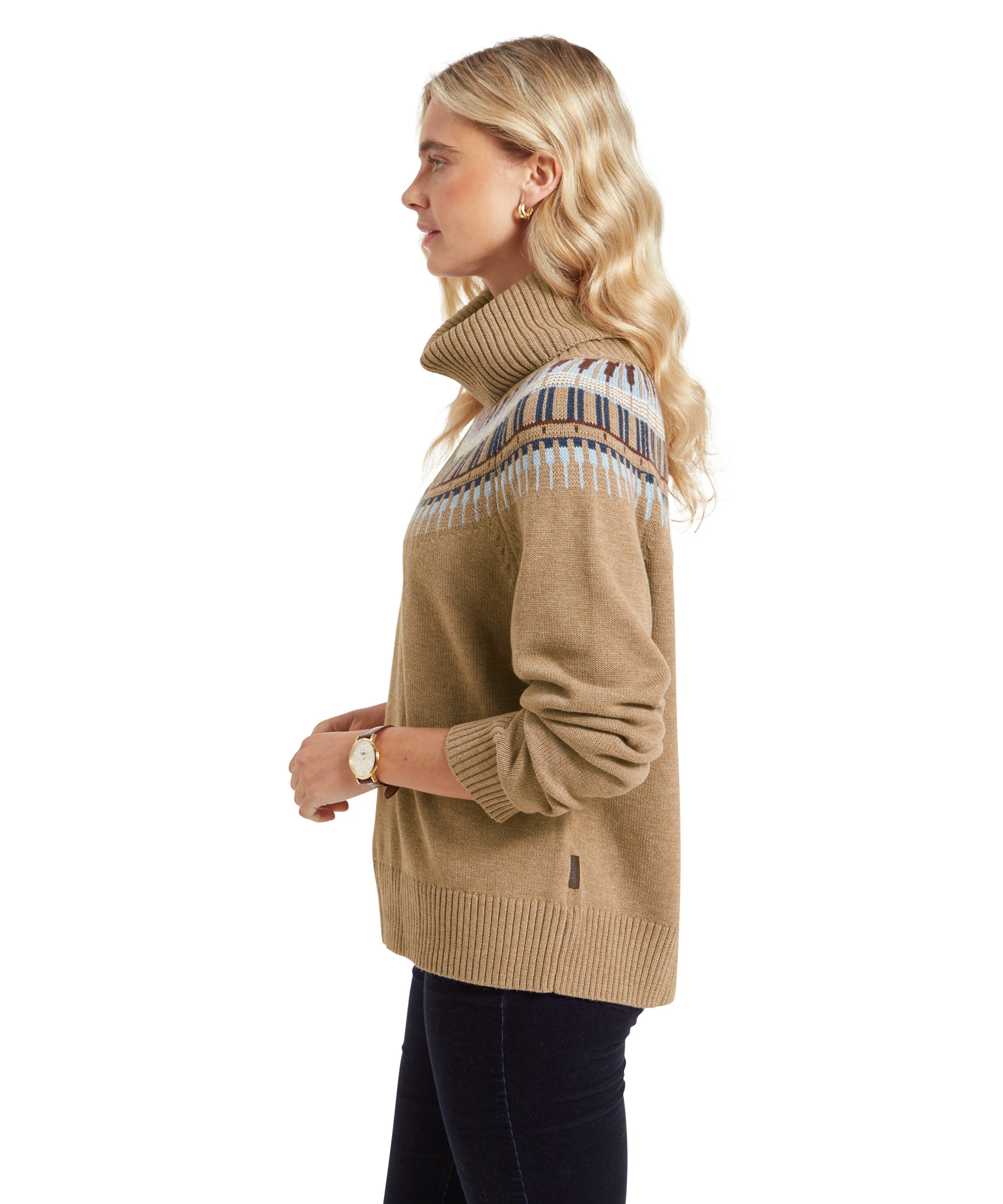 A side profile of the woman in the Schöffel Filey Fairisle Roll Neck Jumper for Women in Beige. She is standing with her arms relaxed, looking slightly forward. The intricate details of the Fair Isle pattern and the ribbed cuffs and hem of the jumper are visible.