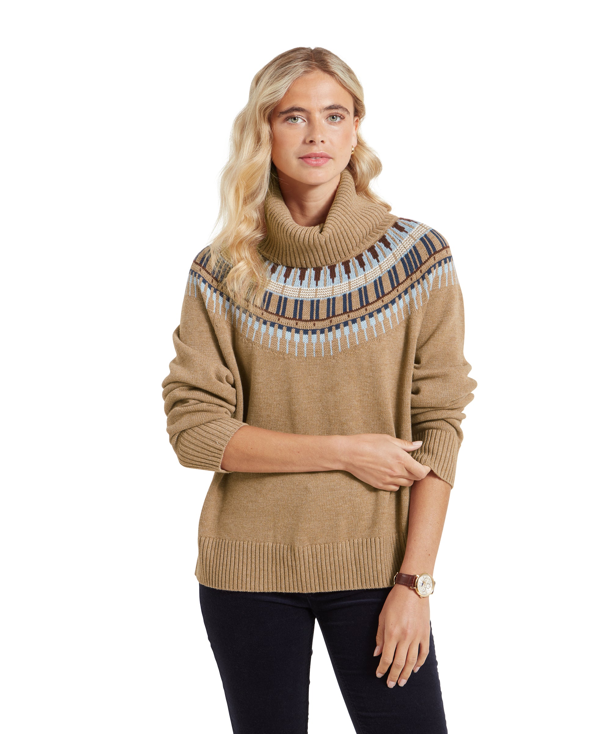 A woman is standing and wearing a Schöffel Filey Fairisle Roll Neck Jumper for Women in Beige with a blue and brown Fair Isle pattern across the shoulders. She has one arm lightly crossed over her midsection, with her other hand resting on her elbow. She is looking directly at the camera with a soft smile. The jumper has a cozy, relaxed fit, paired with dark trousers, and she is wearing a wristwatch on her left hand. Her long, wavy blonde hair is styled down.