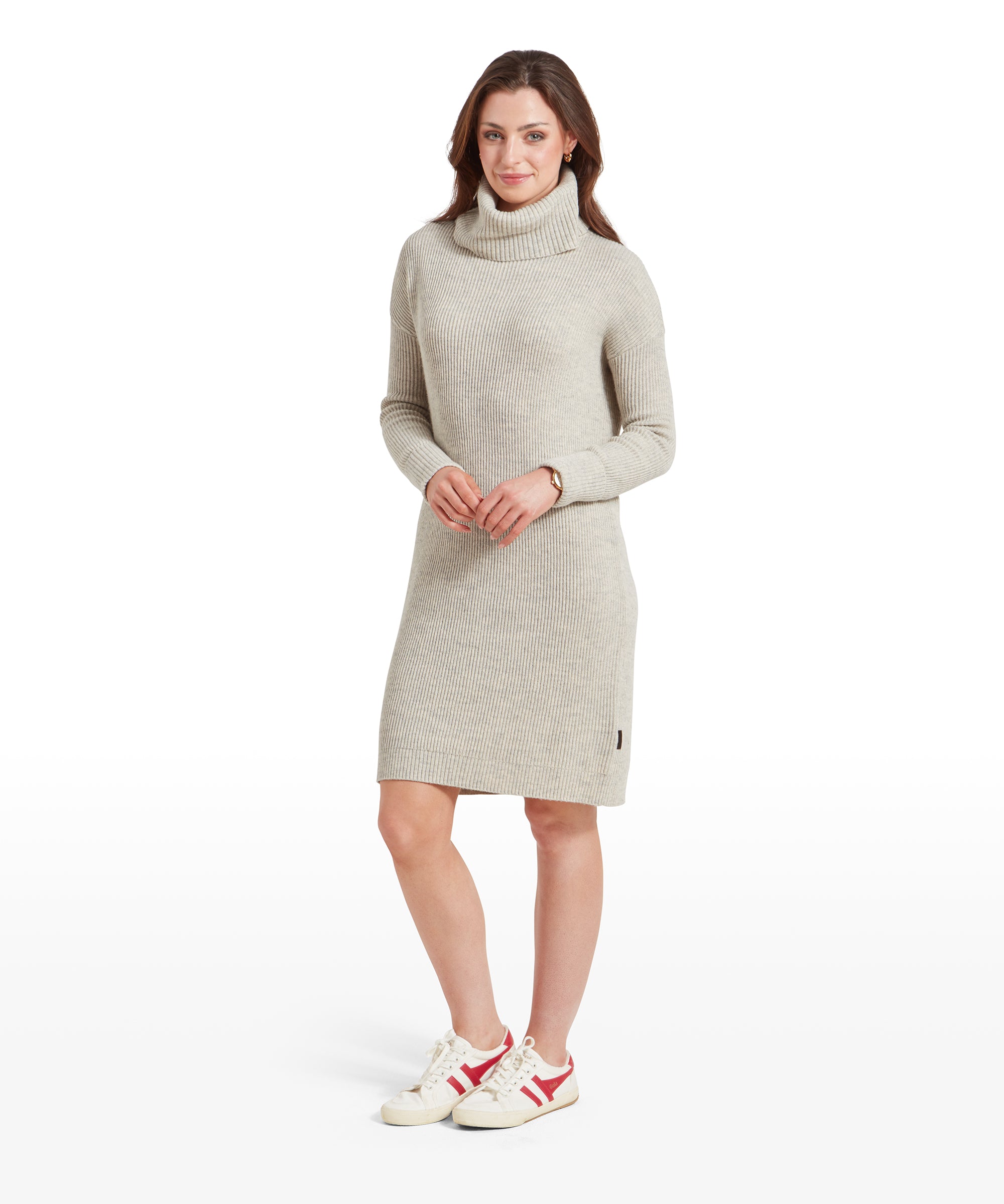 A woman is wearing a Schöffel Filey Roll Neck Dress for Womens in Beige paired with white sneakers with red stripes. She is smiling softly, with her hands clasped in front, and her brown hair falls over her shoulders. The dress is knee-length and fits comfortably.