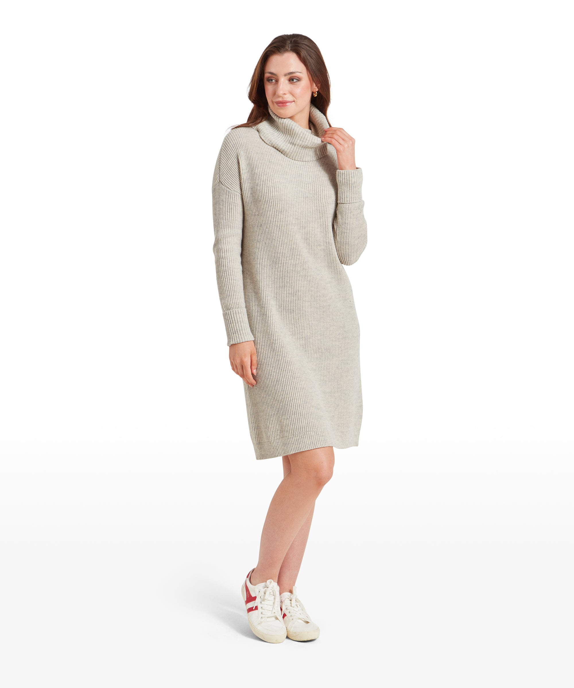 The woman is shown in a Schöffel Filey Roll Neck Dress for Womens in Beige, standing sideways with her head slightly turned forward, looking to her left. Her hands are gently adjusting the roll-neck collar of the dress. She wears casual white trainers with red stripes.