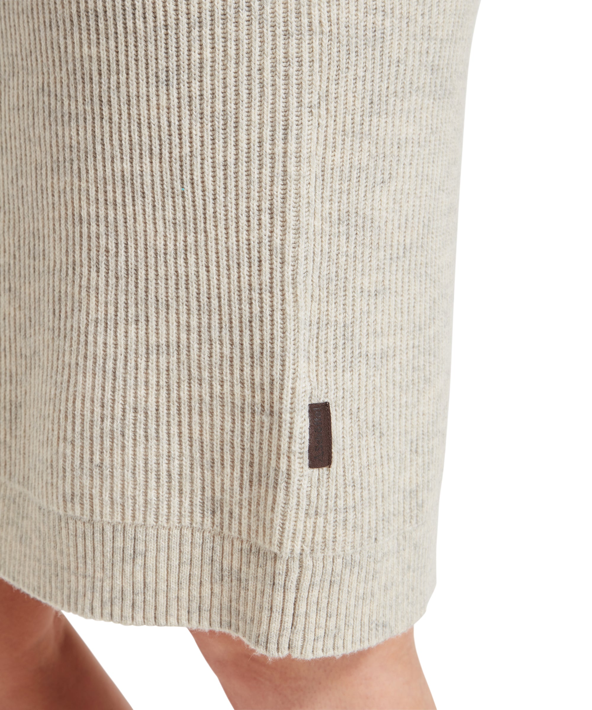 Close-up image showing the hem of a Schöffel Filey Roll Neck Dress for Womens in Beige, revealing a small brown leather tag near the bottom edge of the dress. The fabric appears soft and finely knit.