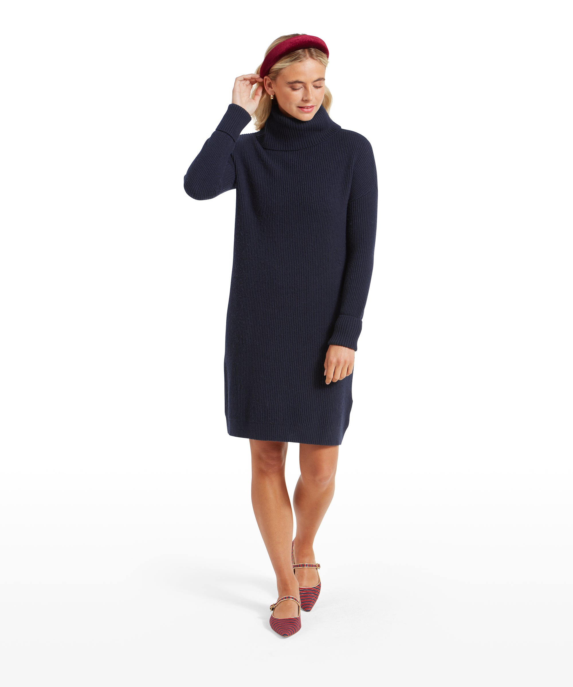 The woman walking forward in the Schöffel Filey Roll Neck Dress for Womens in Navy, adjusting her hair with her hand, wearing the same burgundy headband and red striped flats.