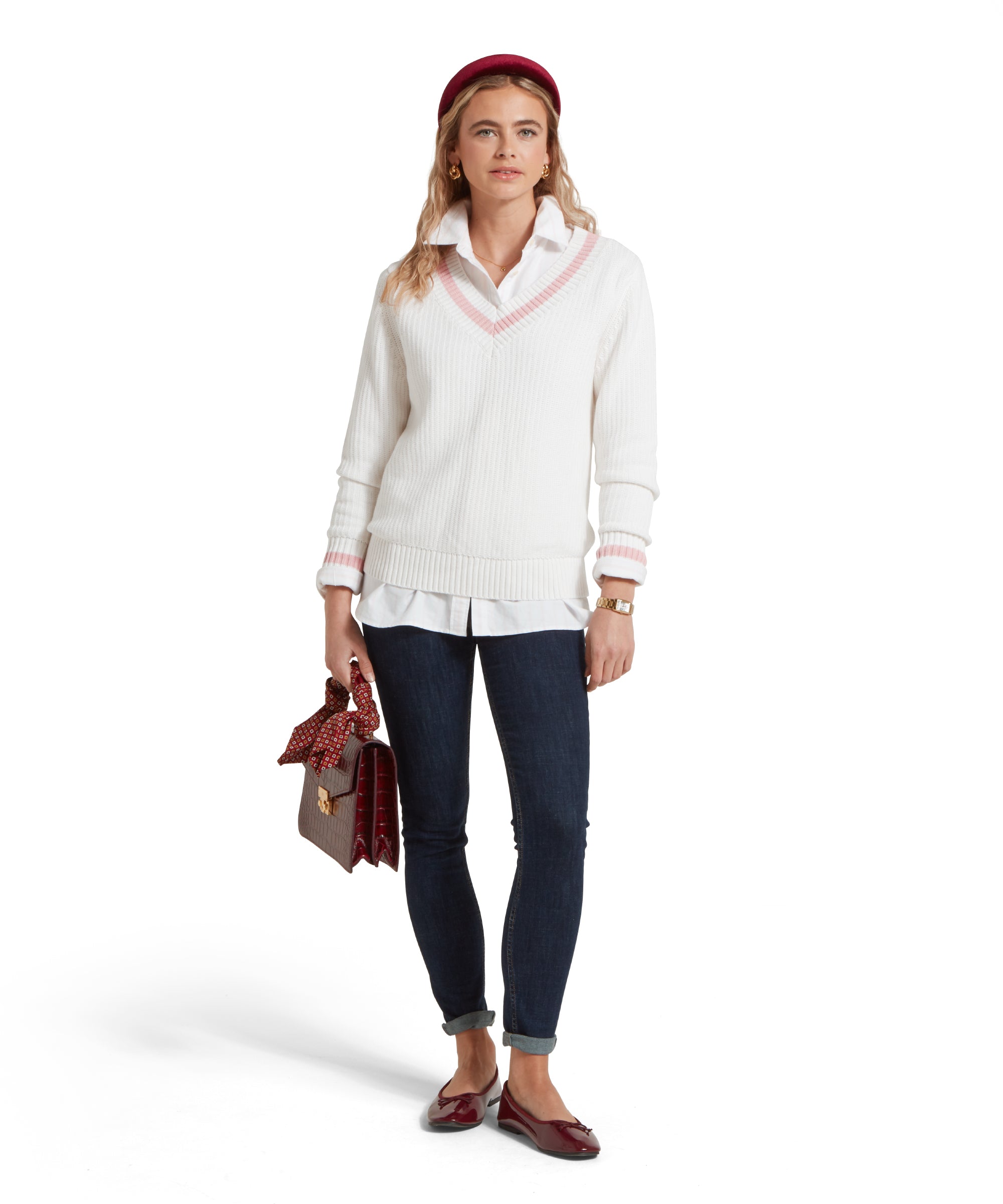 A full-height image of a female model wearing a white Schöffel jumper with pink trim on V-neck collar and cuffs, paired with a white shirt, dark denim jeans and dark red pumps.