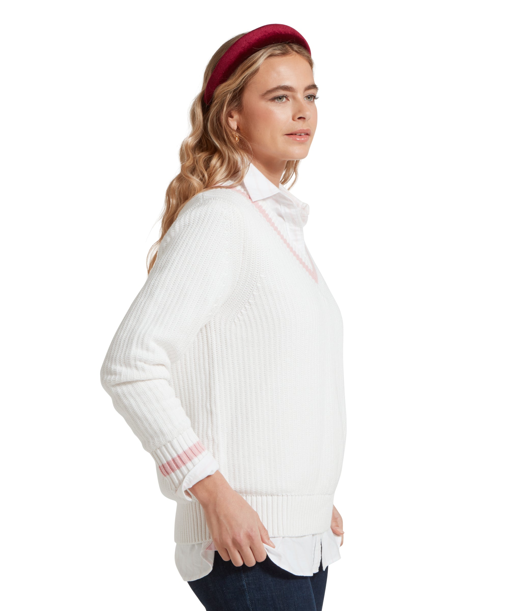 A side view image of a female model wearing a white Schöffel jumper with pink trim on V-neck collar and cuffs, paired with a white shirt, dark denim jeans and red headband.