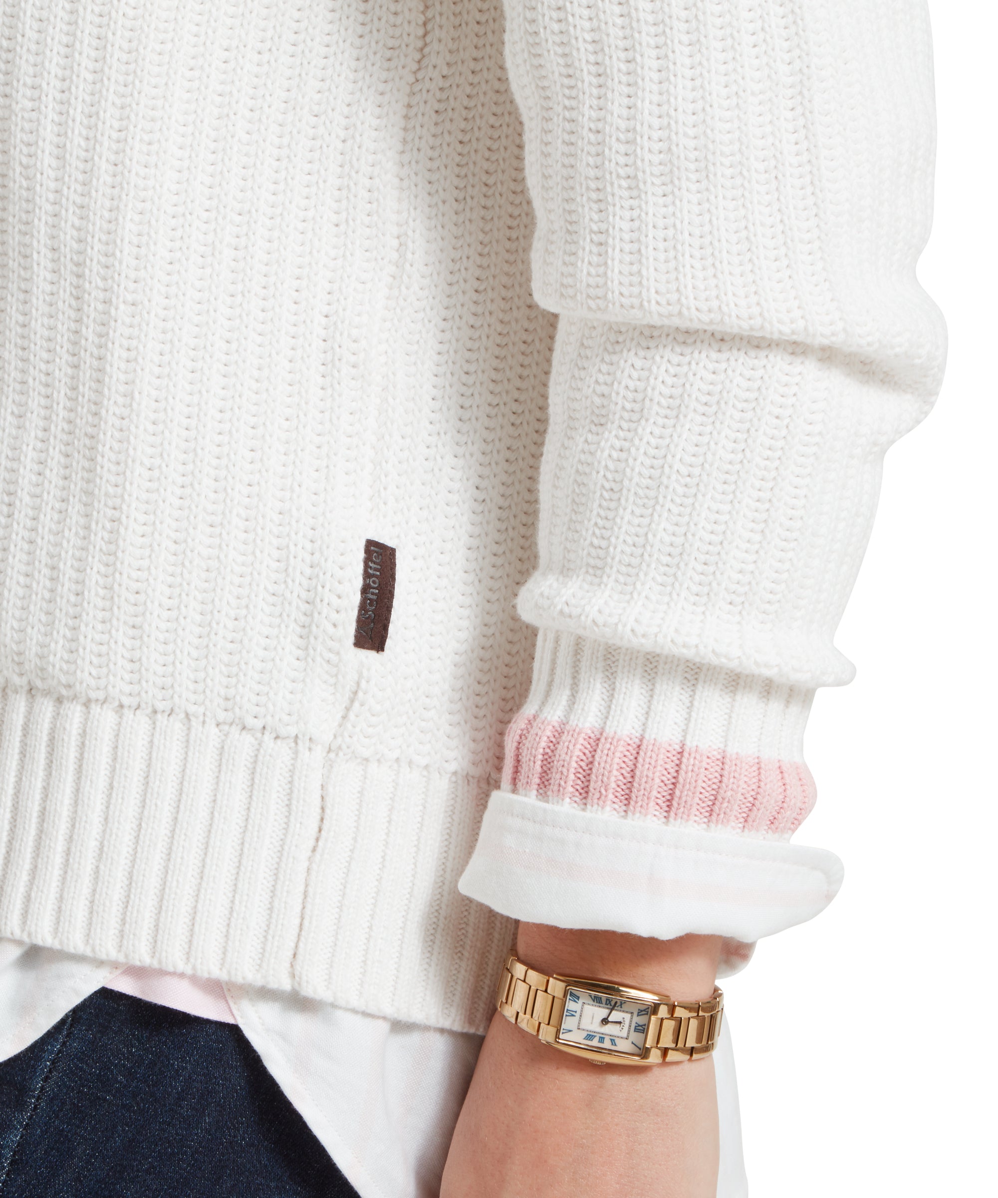 A close up image of cuffs and hem of a white Schöffel jumper with pink trim on cuffs, paired with a white shirt, dark denim jeans and gold accessories.