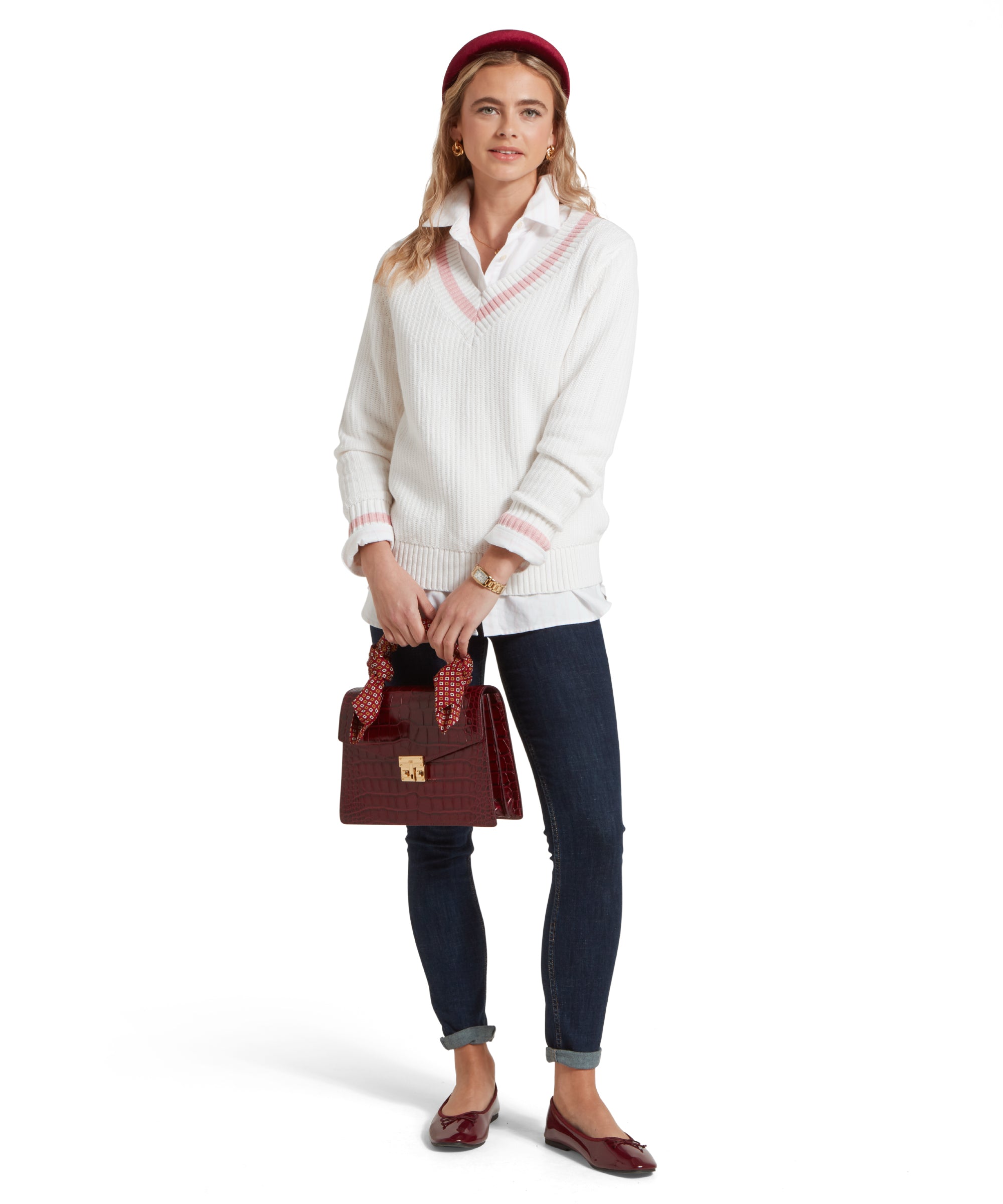 A full height image of a female model wearing a white Schöffel jumper with pink trim on V-neck collar and cuffs, paired with a white shirt, dark denim jeans and red pumps. She is standing in a relaxed pose holding a dark red leather handbag.