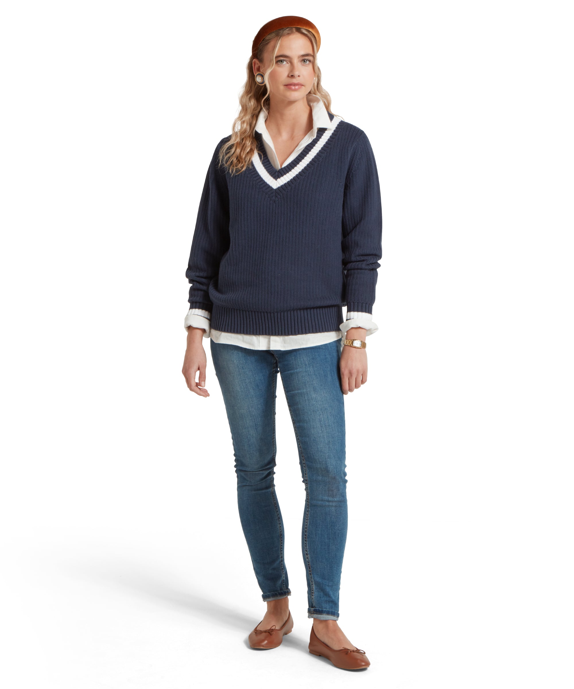 A full height image of a female model wearing a navy Schöffel jumper with white trim on V-neck collar and cuffs, paired with a white shirt, mid denim jeans, tan pumps and gold accessories.