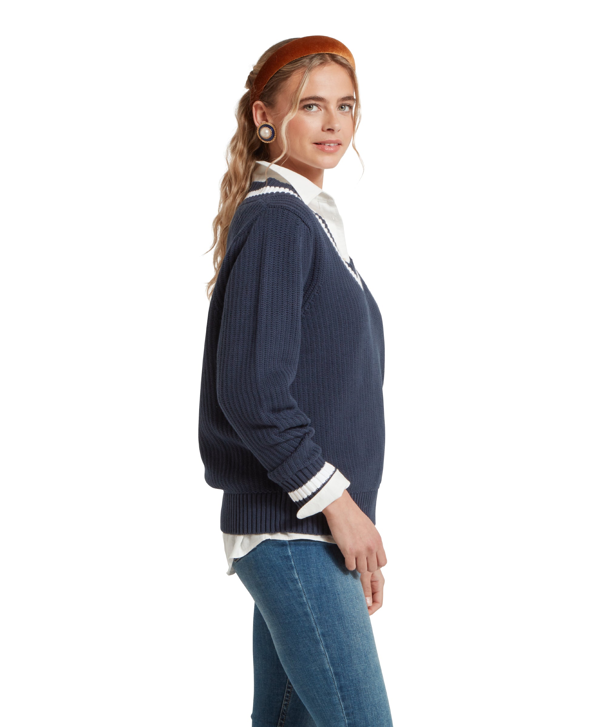 A side view image of a female model wearing a navy Schöffel jumper with white trim on V-neck collar and cuffs, paired with a white shirt, mid denim jeans and gold accessories.