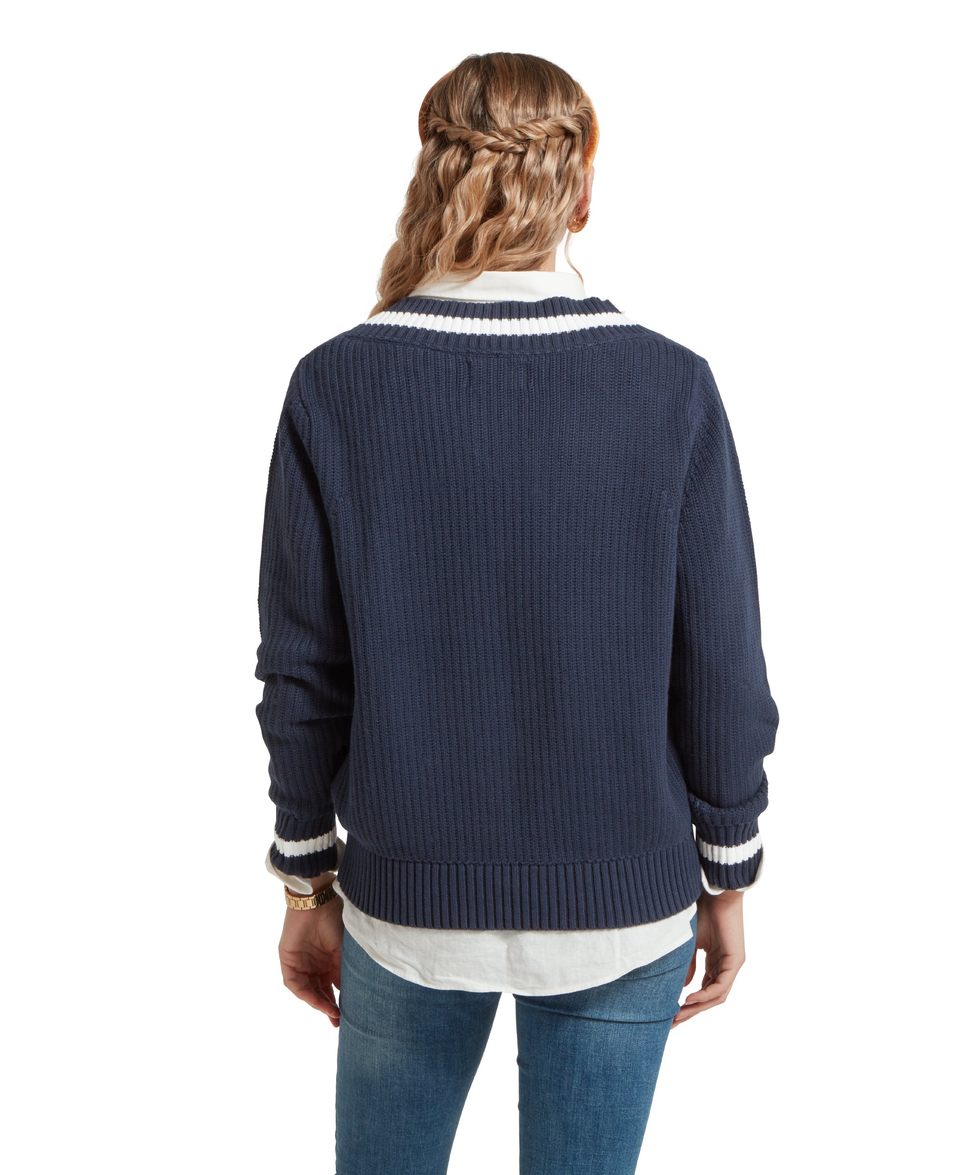 A back view image of a female model wearing a navy Schöffel jumper with white trim on V-neck collar and cuffs, paired with a white shirt, mid denim jeans and gold accessories.