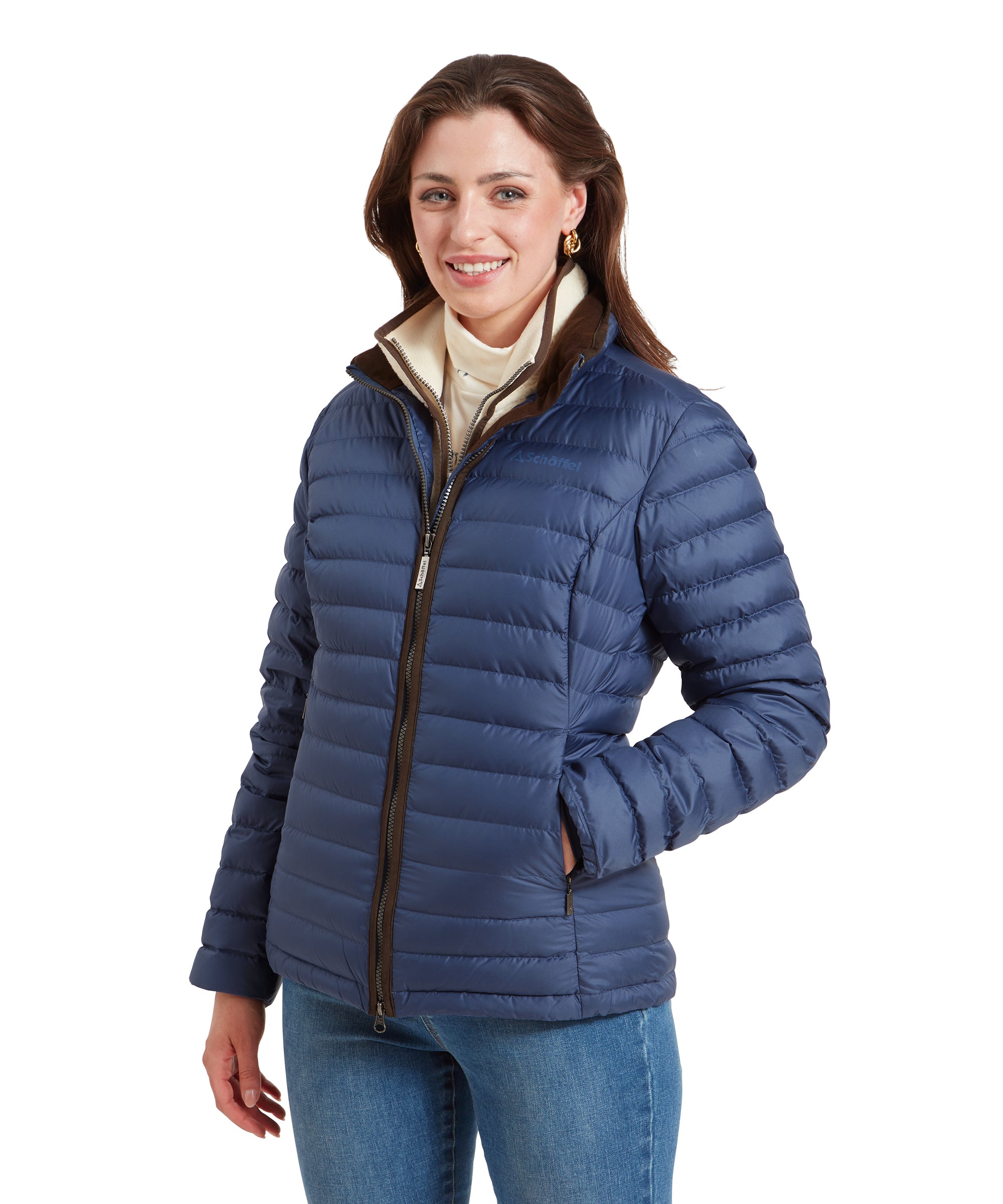 A woman wearing a Schöffel Hambleton Down Jacket for Women in Blue with a brown inner collar. The jacket is zipped up, and she has her hands in her pockets. She is smiling and looking straight ahead.