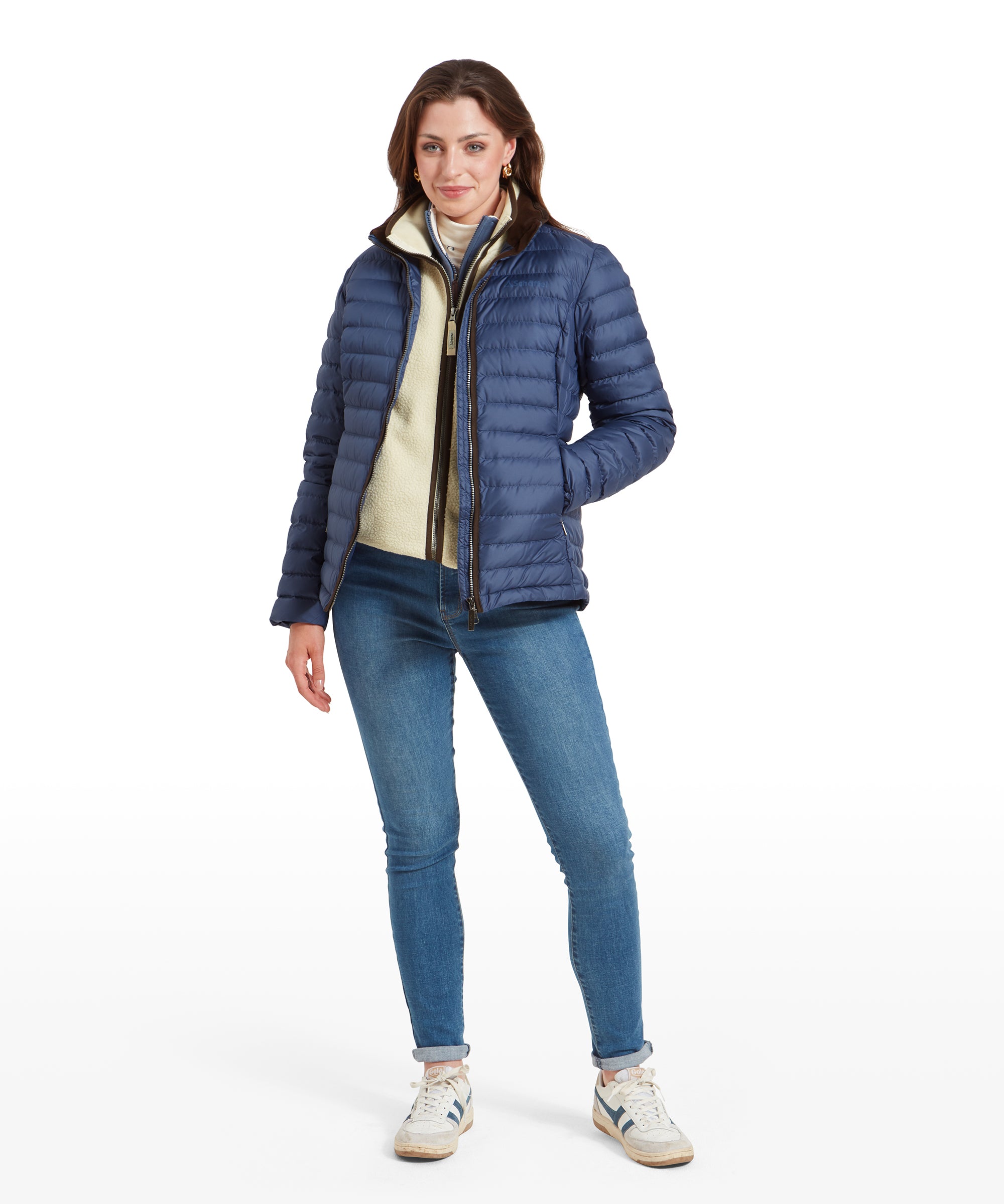 A woman wearing a Schöffel Hambleton Down Jacket for Women in Blue over a cream-colored sweater. The jacket is partially unzipped, revealing the sweater underneath. She is also wearing blue jeans and white trainers, standing with one hand in her pocket.