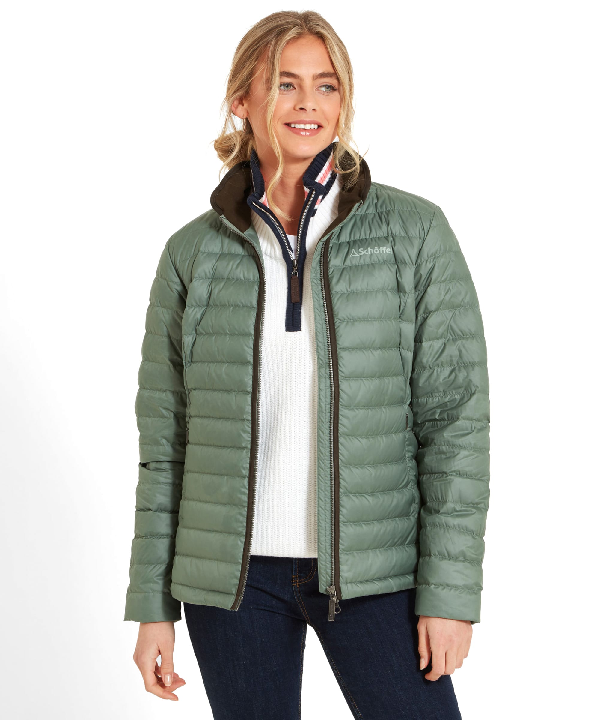 A woman wearing a Schöffel Hambleton Down Jacket for Women in Green over a white sweater. She is smiling, and her blonde hair is styled in a casual updo. The jacket has a high collar with brown trim, and she is posing with her hands relaxed at her sides.