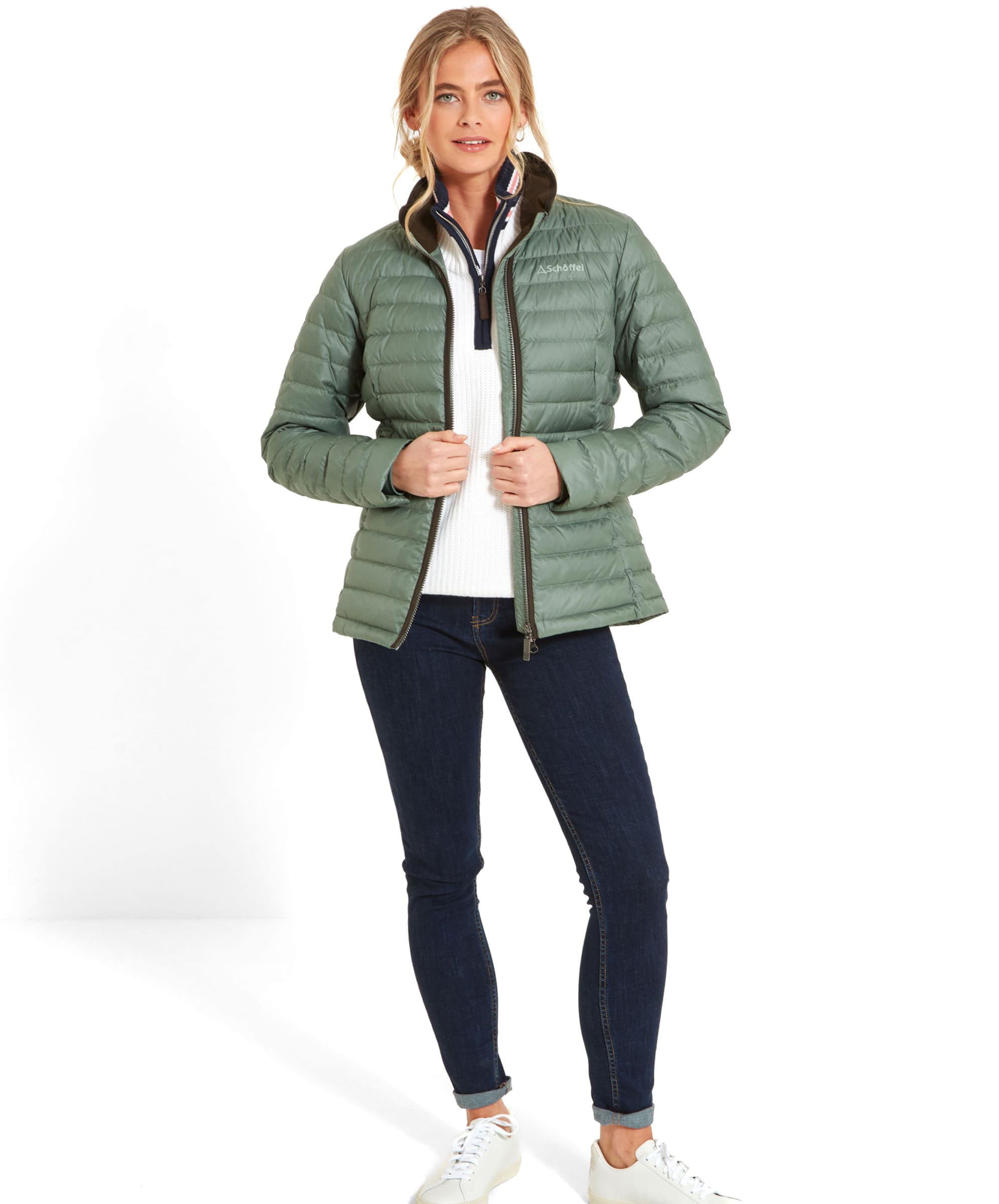 A full-body image of a woman dressed in a Schöffel Hambleton Down Jacket for Women in Green, white sweater, dark jeans, and white trainers. She is smiling and standing with her hands lightly grasping the sides of her jacket, as if zipping it up.