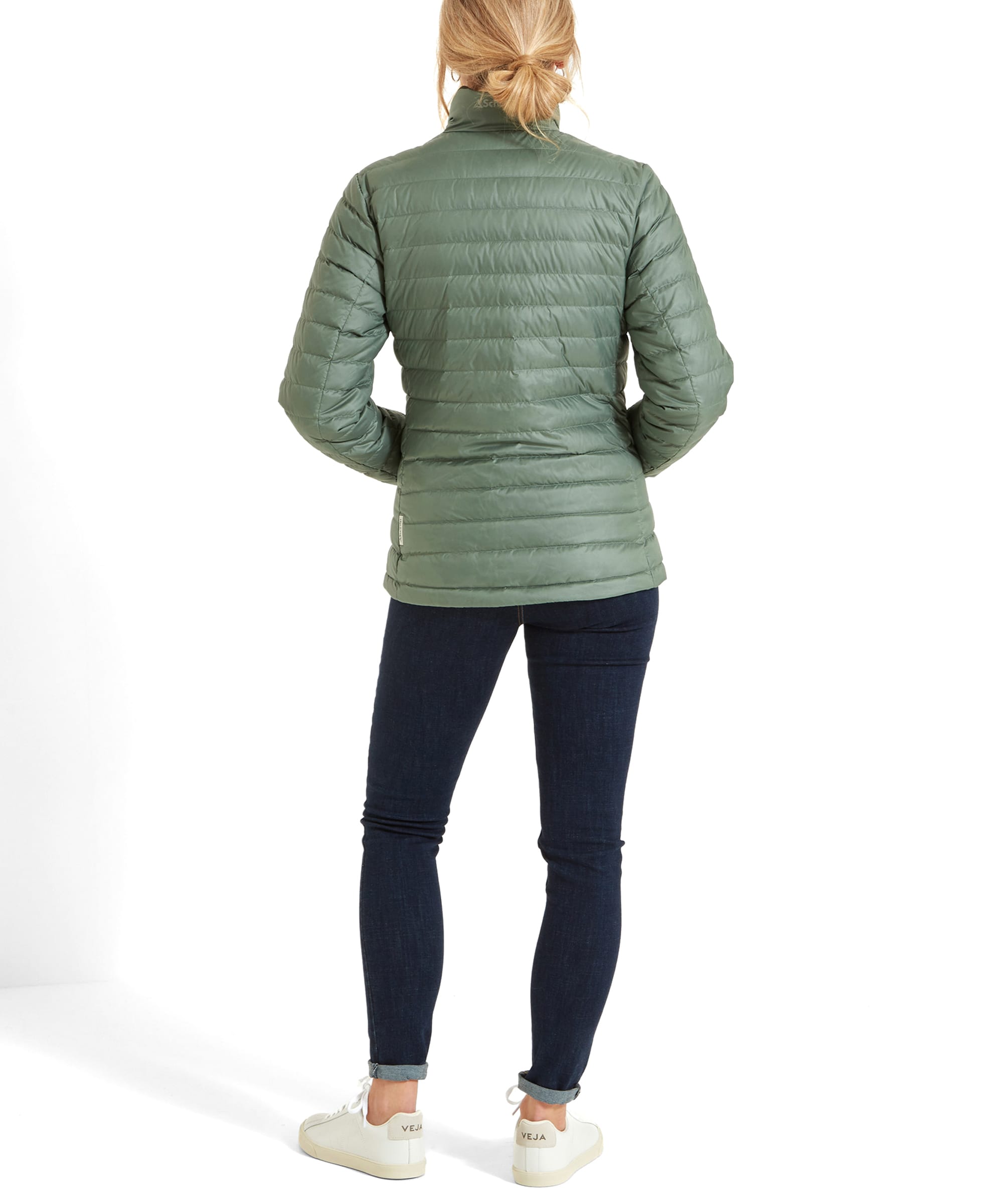 Rear view of a woman wearing a Schöffel Hambleton Down Jacket for Women in Green, showing the jacket's back design. She is also wearing dark jeans and white sneakers, with her blonde hair tied in a low ponytail.