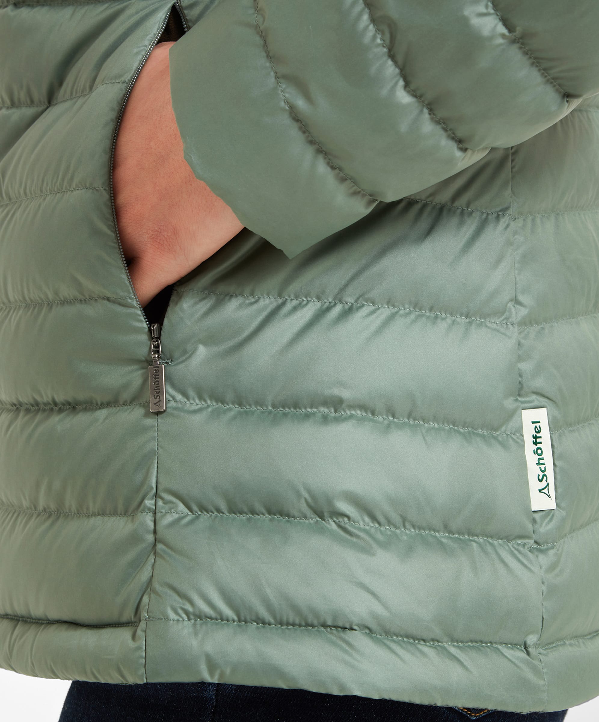 Close-up of a hand reaching into the pocket of a Schöffel Hambleton Down Jacket for Women in Green. The focus is on the pocket zipper, which is partially open, showing the detailed stitching and the Schöffel logo tag on the jacket's hem.